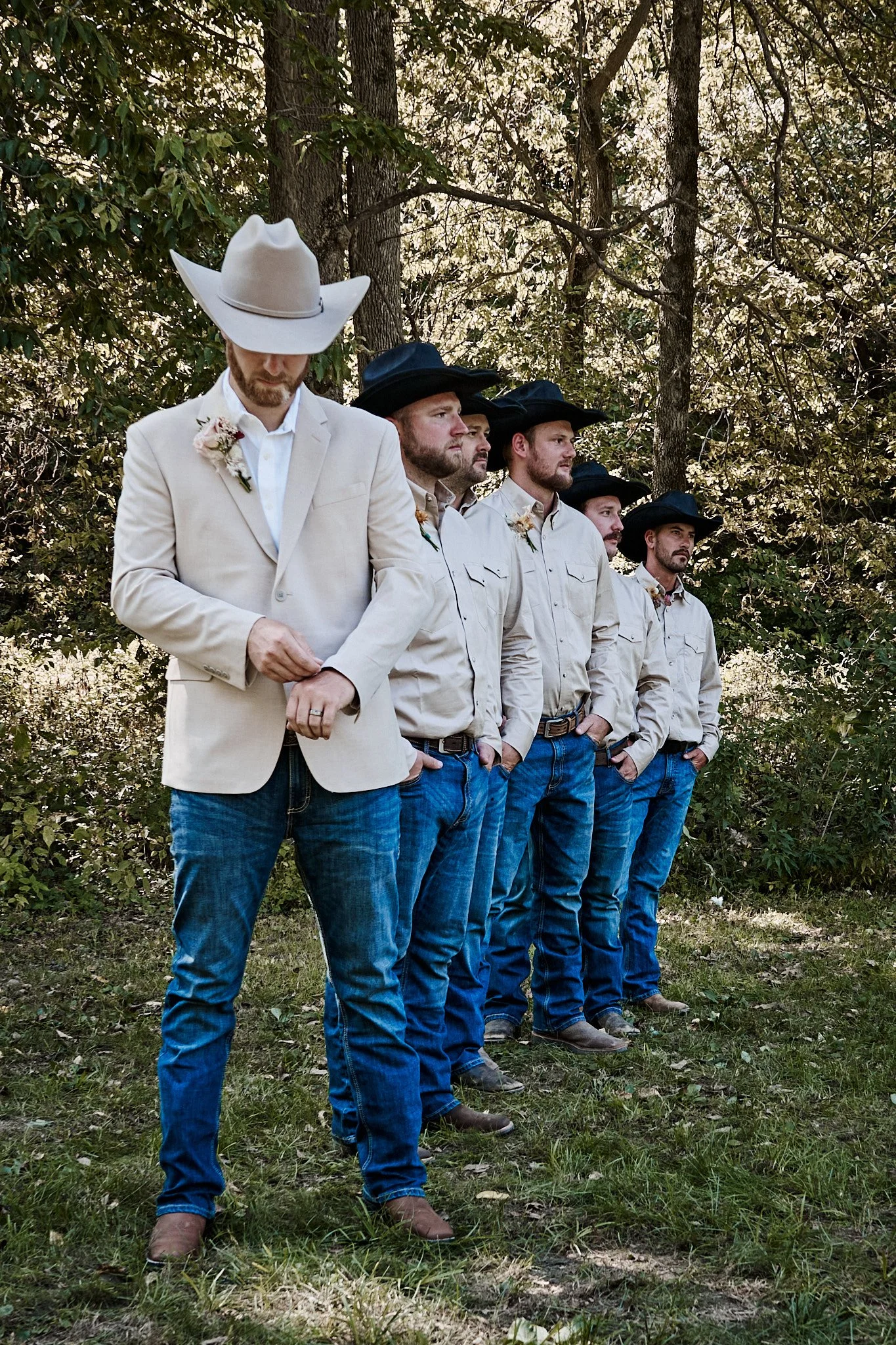 Five men standing in a line outdoors among trees, all wearing cowboy hats and western attire, with the man at the front looking down.