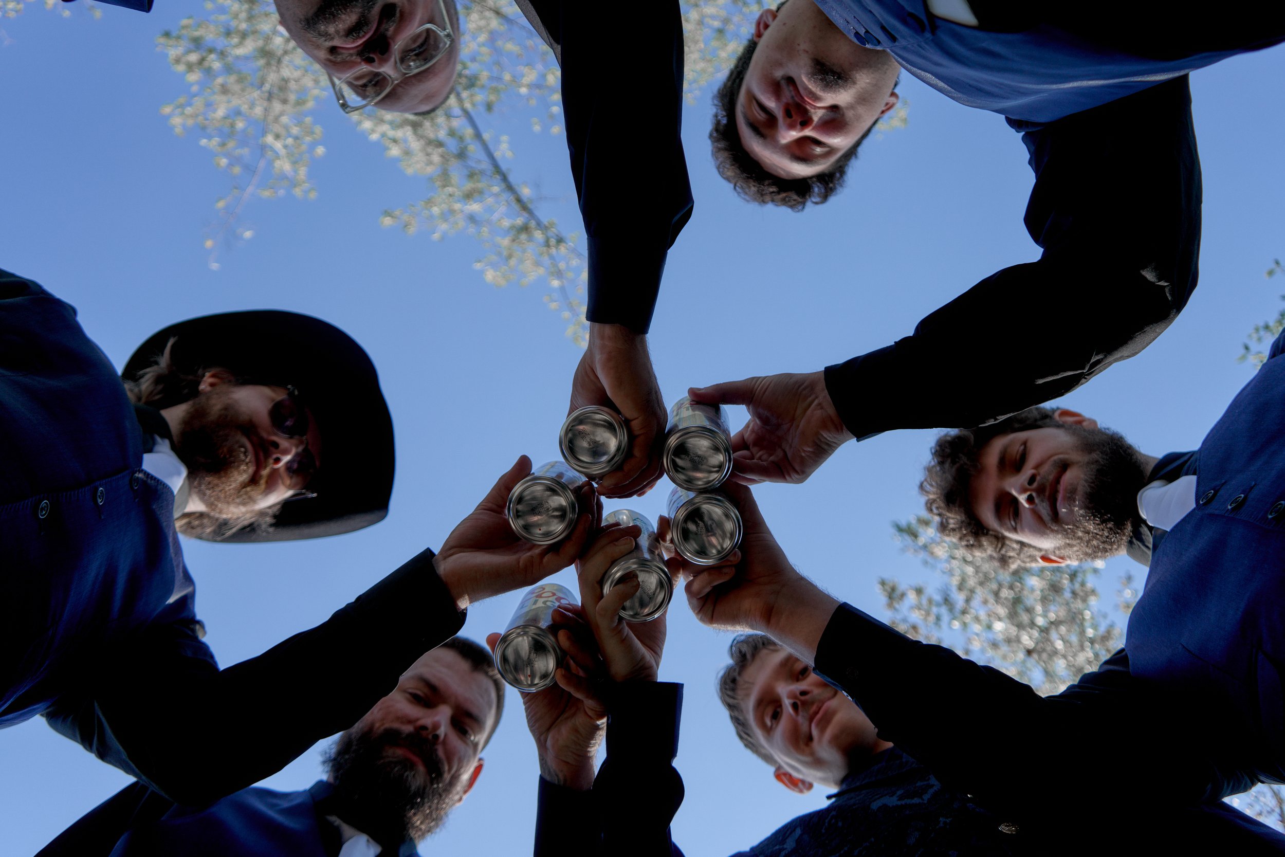 A group of people with varying ages and ethnicities standing in a circle outdoors, holding cans together in a toast. The photo is taken from below against a clear blue sky with some tree branches visible.