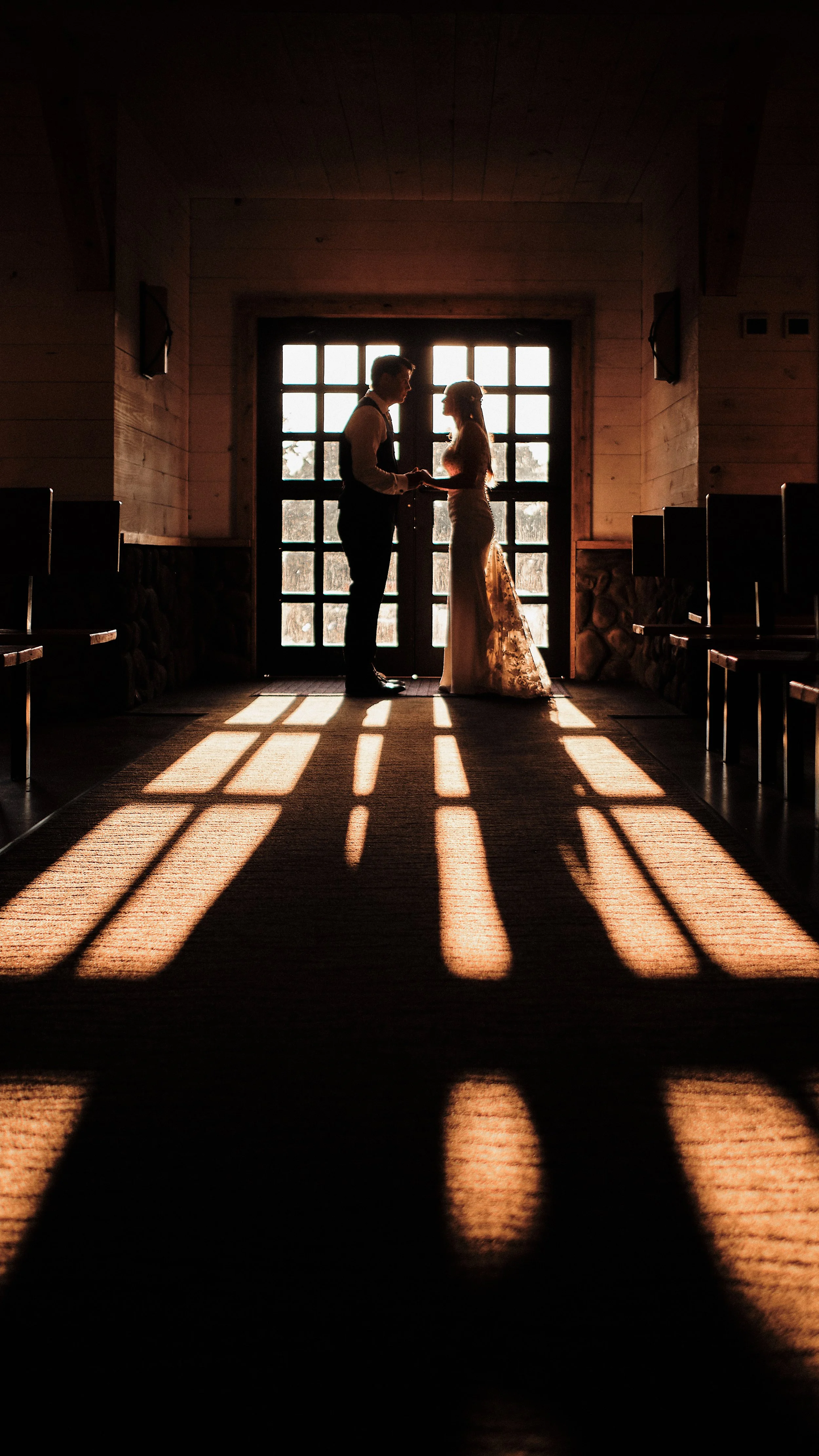 A silhouette of a couple holding hands and looking at each other in front of a large window with sunlight streaming through, casting shadows on the floor inside a rustic wooden building.
