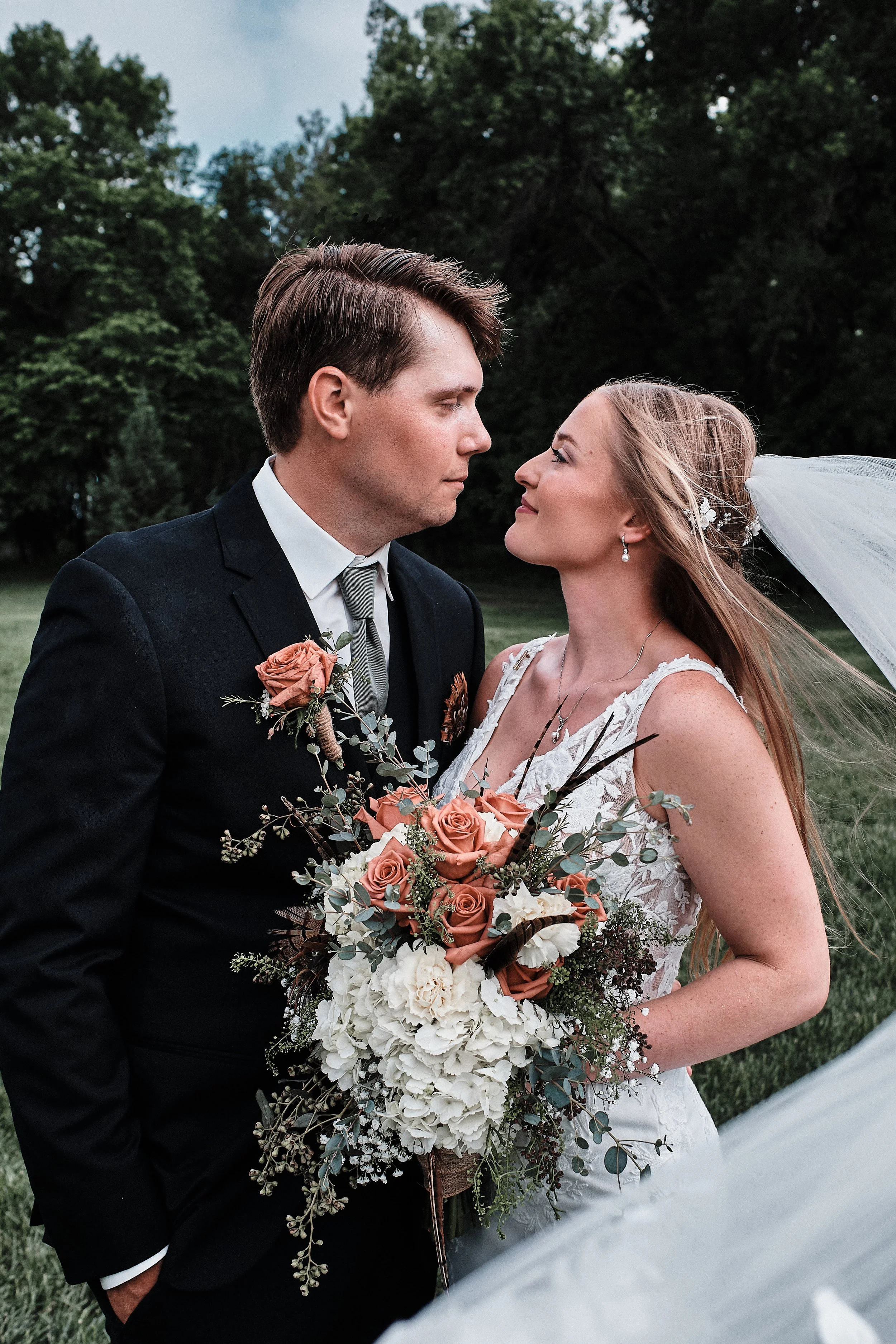 A bride and groom at their wedding, facing each other outdoors with greenery in the background. The bride is holding a large bouquet of flowers, and the groom has a boutonniere.