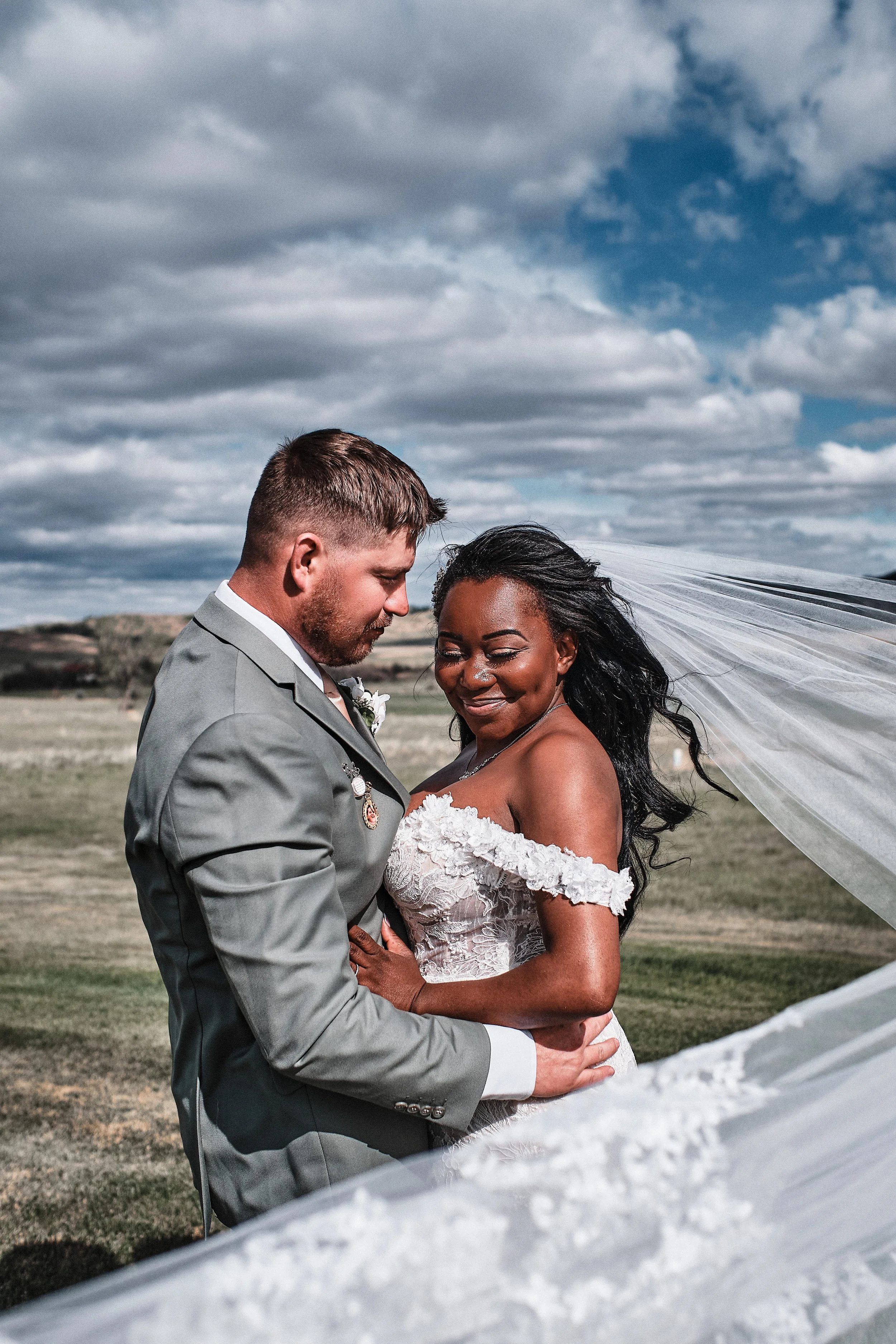 A newlywed couple stands close together outdoors under a cloudy sky, smiling with eyes closed, sharing an intimate moment. The groom is dressed in a gray suit with a white shirt, and the bride wears an off-the-shoulder lace wedding dress. Her long bl