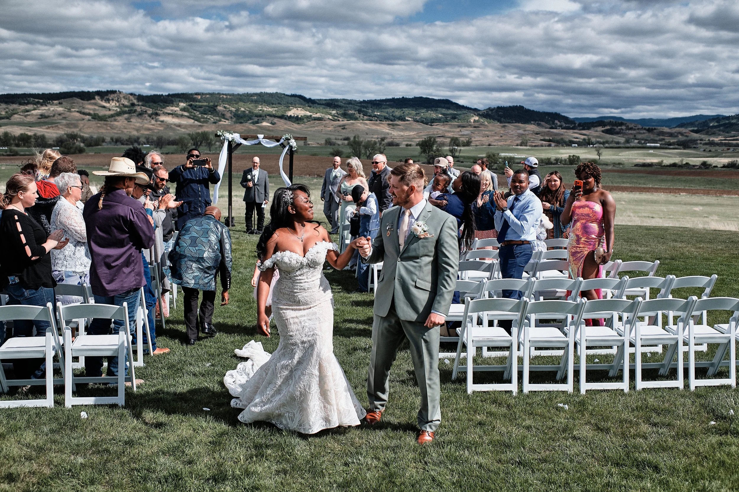 A bride and groom walking hand in hand after their wedding ceremony outdoors with guests seated and standing around them. The scene is set in a green field with a backdrop of rolling hills and a cloudy sky.