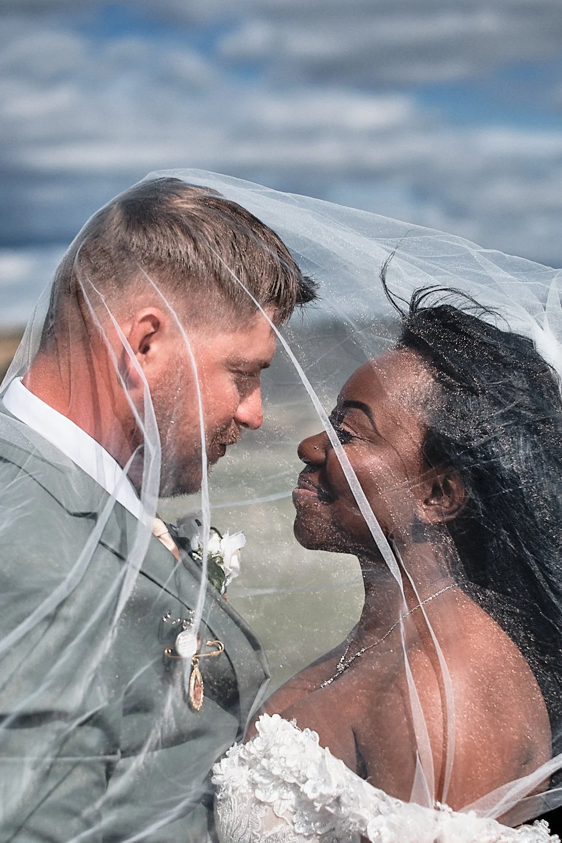 A newlywed couple on their wedding day, standing close with their foreheads touching, under a sheer veil, at the beach with ocean and sky in the background.