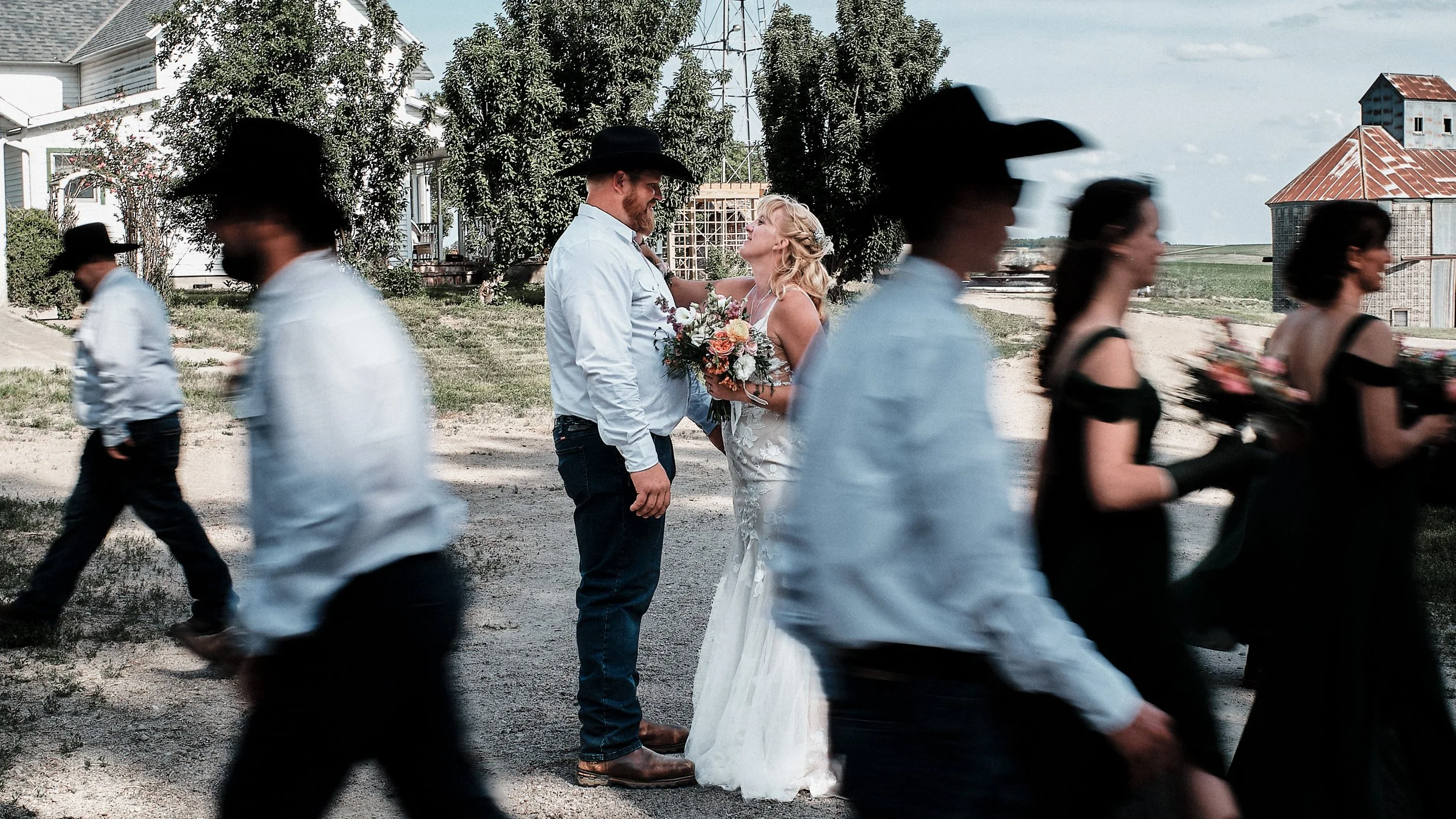 A couple in cowboy hats and Western attire sharing a moment amidst a line of moving wedding guests in outdoor setting with trees and farm buildings.