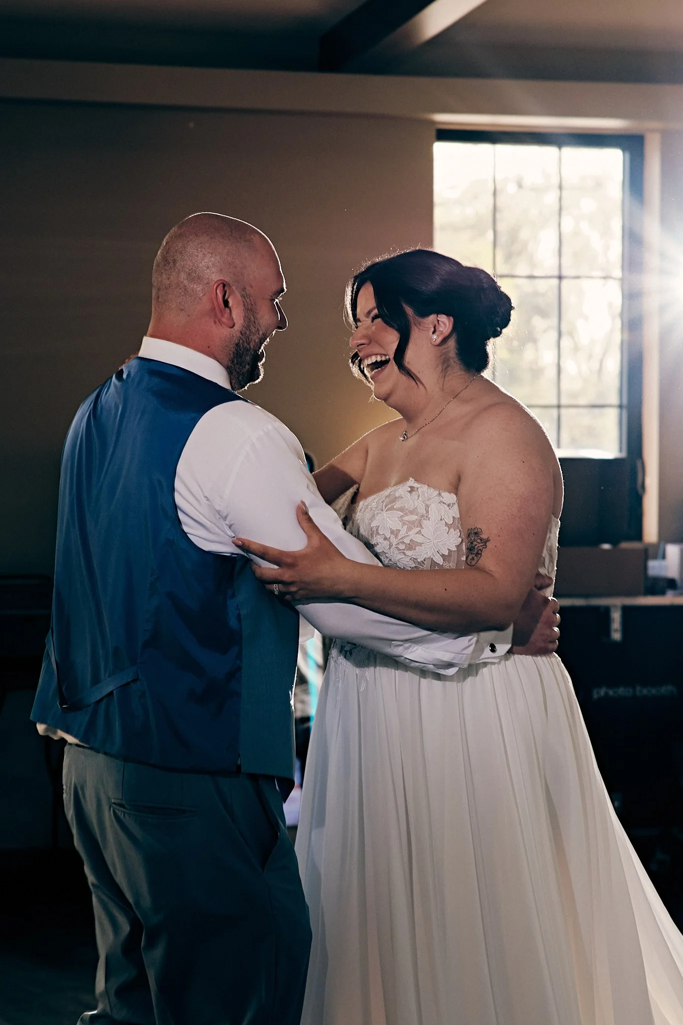 A bride and groom sharing a dance indoors with natural light coming through a window, both smiling and looking at each other.