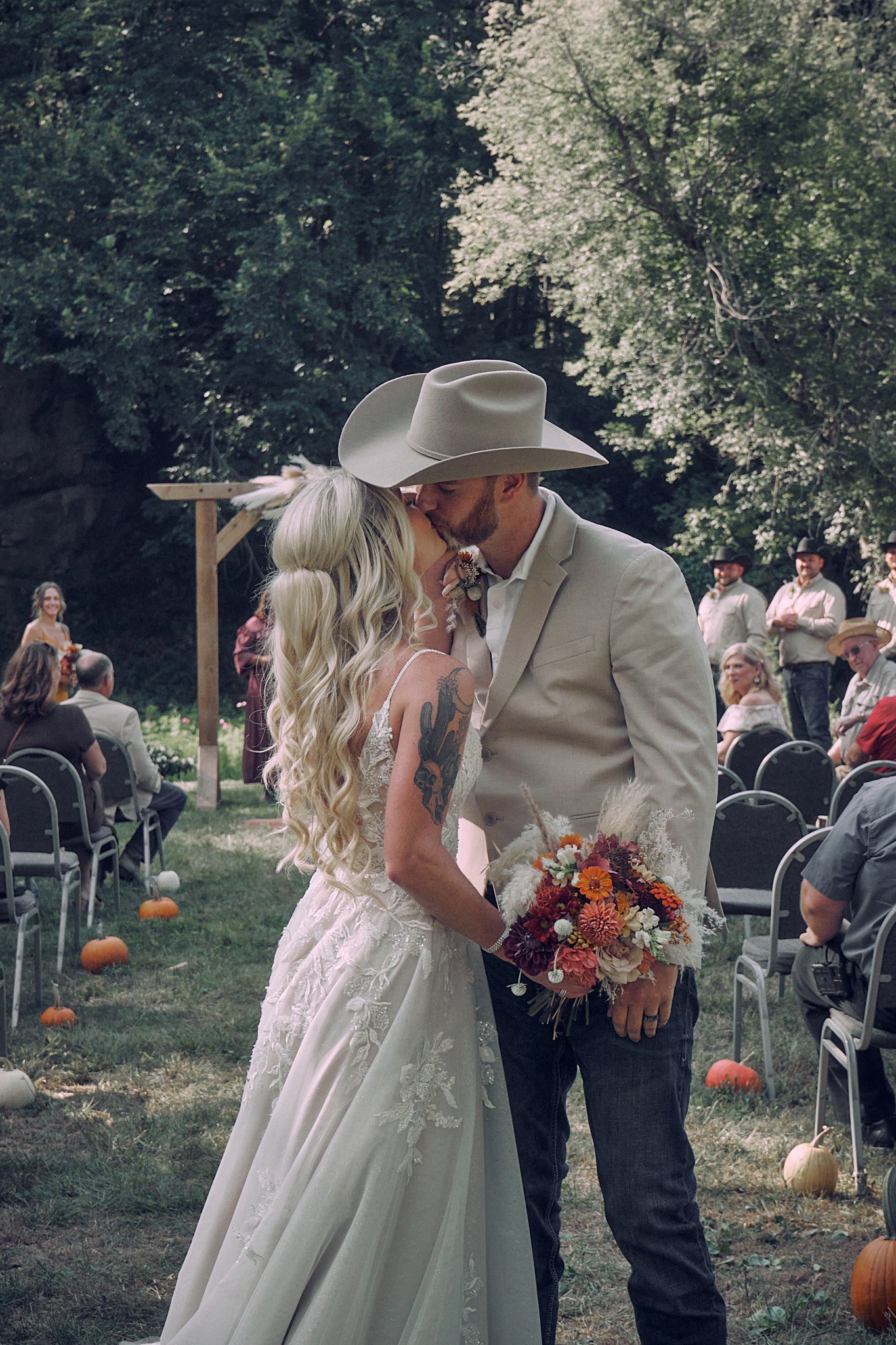 A couple is kissing at an outdoor wedding ceremony, with guests seated in chairs behind them. The bride has long, wavy blonde hair and tattoos on her arm, wearing a white wedding gown. The groom has a beard, wearing a beige suit and cowboy hat. The s
