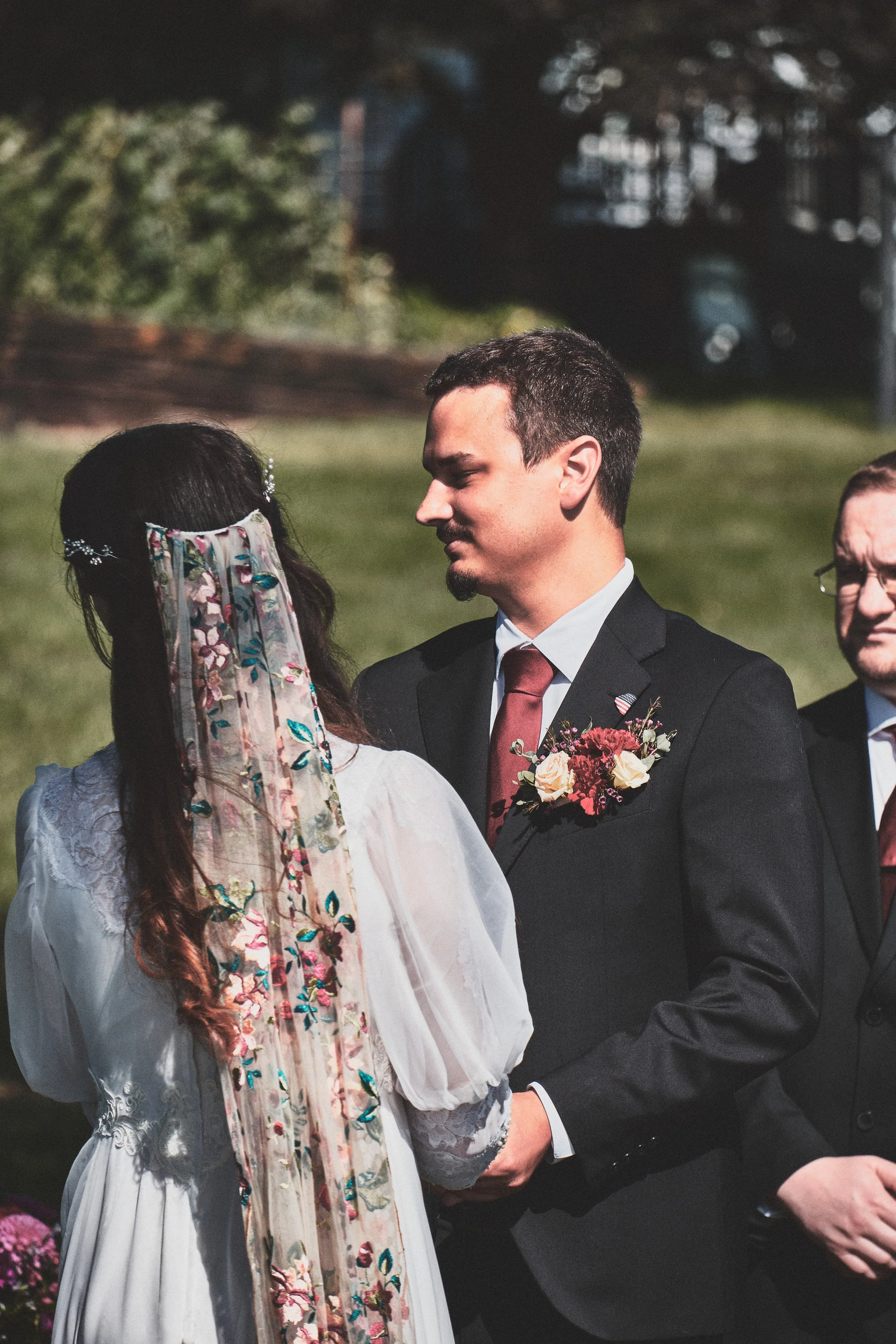 A couple holding hands during a wedding ceremony outdoors, with a woman in a white dress with floral embroidery and a man in a black suit with a tie and boutonniere, standing on a grassy area with trees in the background.