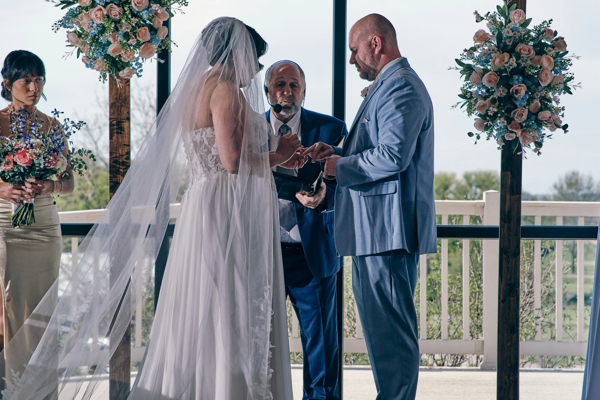 A couple exchange wedding rings during their outdoor wedding ceremony, with an officiant and bridesmaid present, surrounded by large floral arrangements.