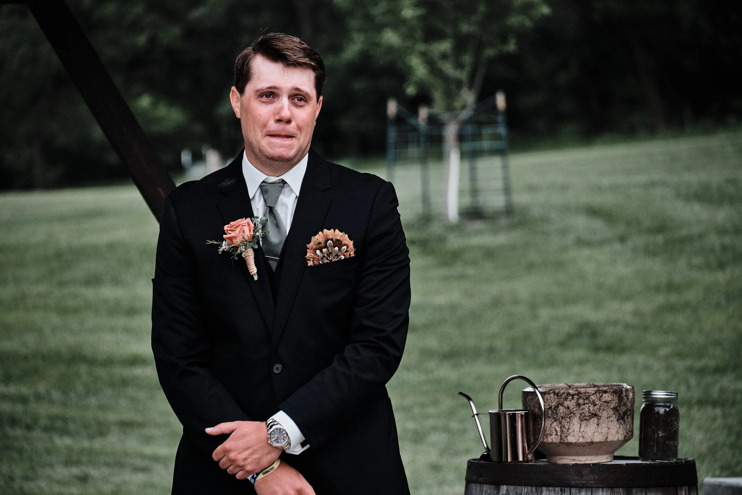A man in a black suit and tie standing outdoors during daytime, with a grassy field and trees in the background. He appears emotional, with tears in his eyes, and has a boutonniere on his lapel. There is a table next to him with a metal teapot and ot
