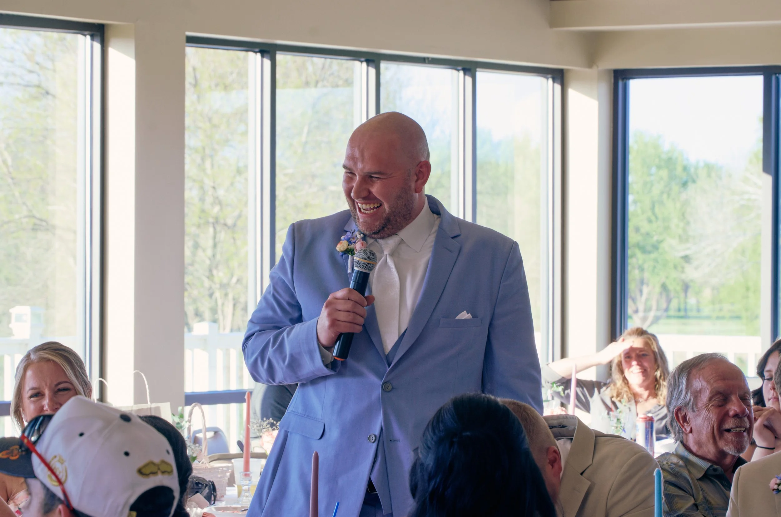 A man in a light blue suit and white shirt is speaking into a microphone at a social gathering, smiling. He is bald and has a boutonniere on his lapel. Several people are seated at tables, and large windows reveal a green outdoor scene.