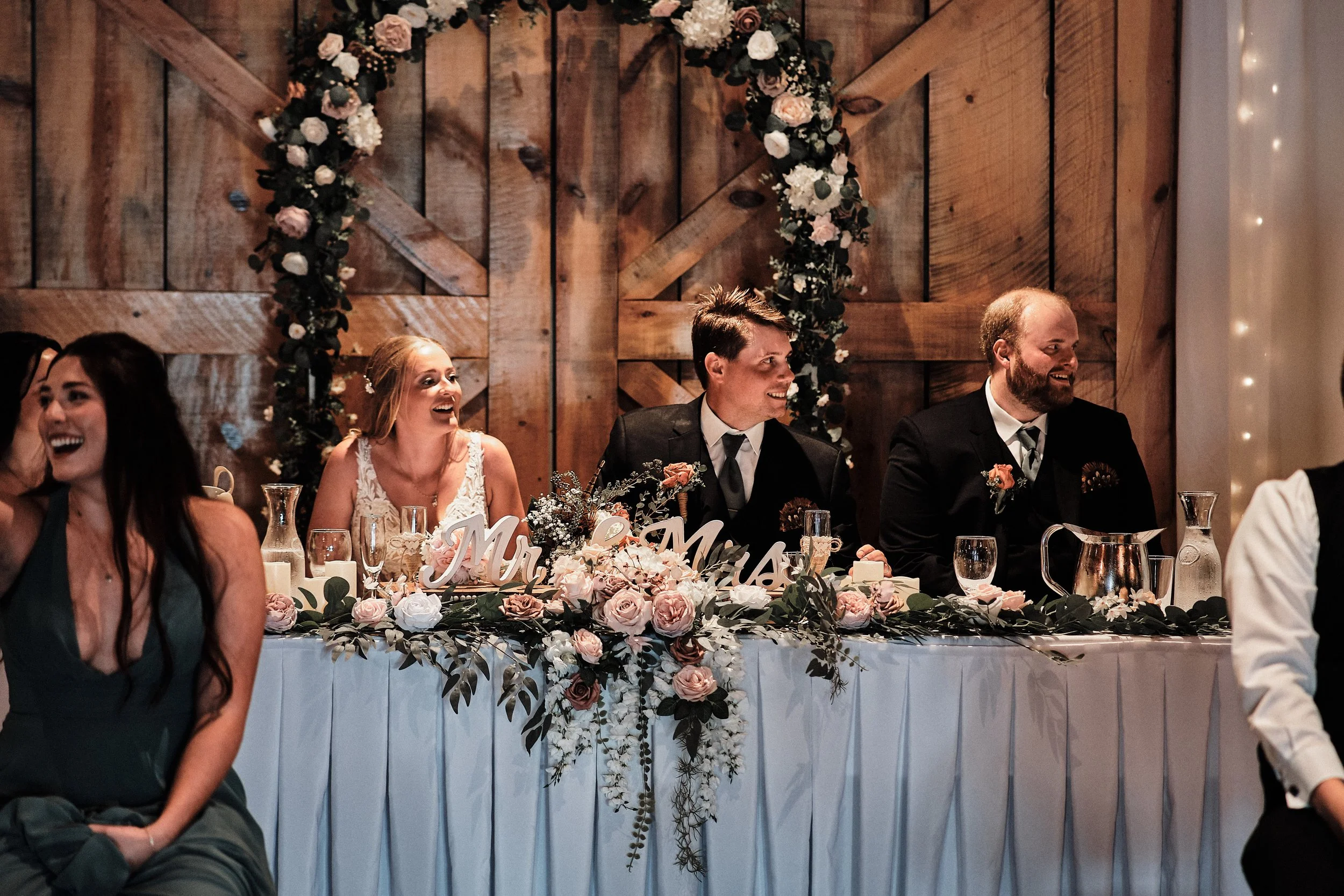 People sitting at a wedding reception table decorated with pink and white flowers. In the background, a wooden wall and floral arch are visible. The bride and groom are smiling in the center of the image, dressed in formal attire.