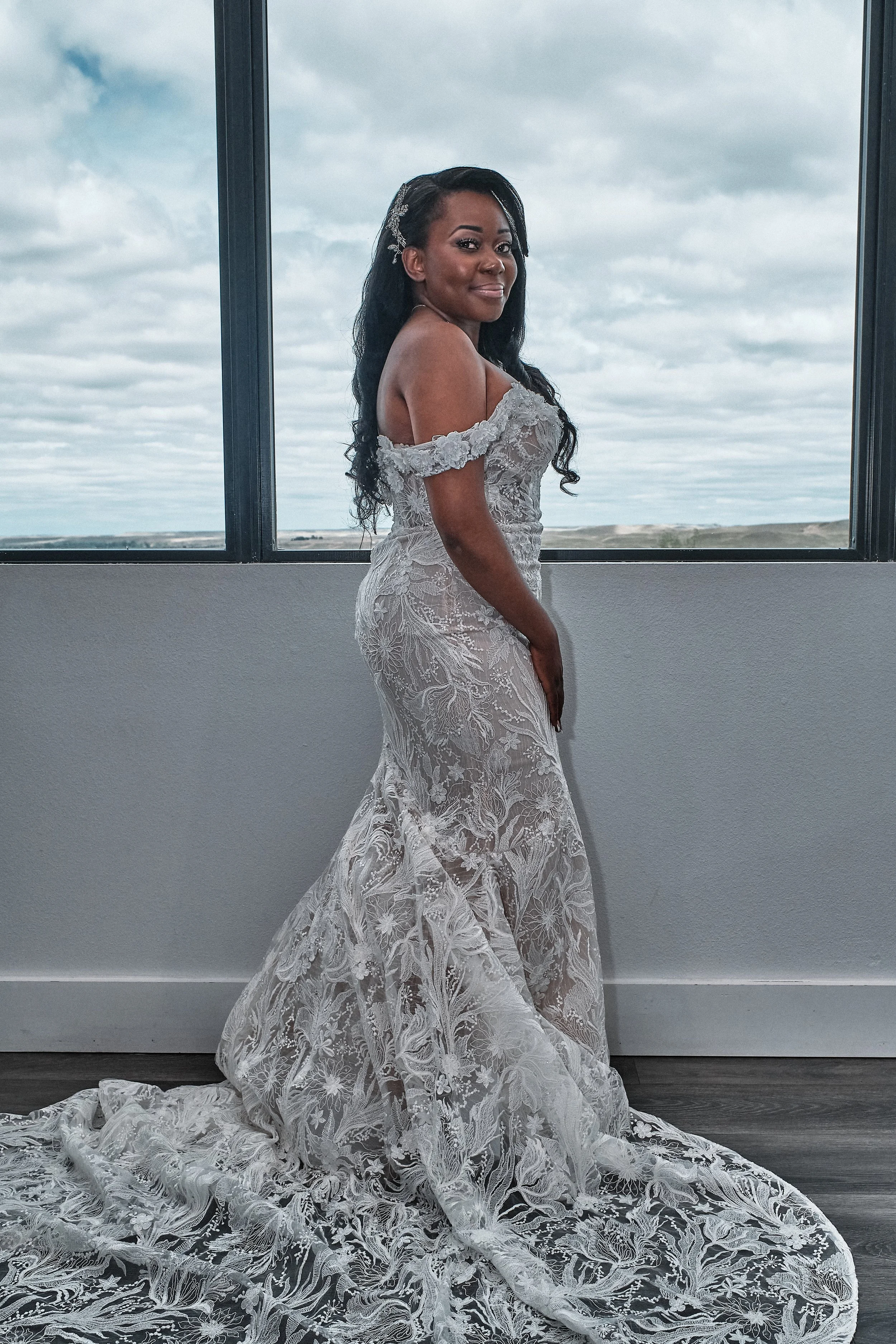 A woman in a white lace wedding dress standing indoors near a large window with a cloudy sky in the background.