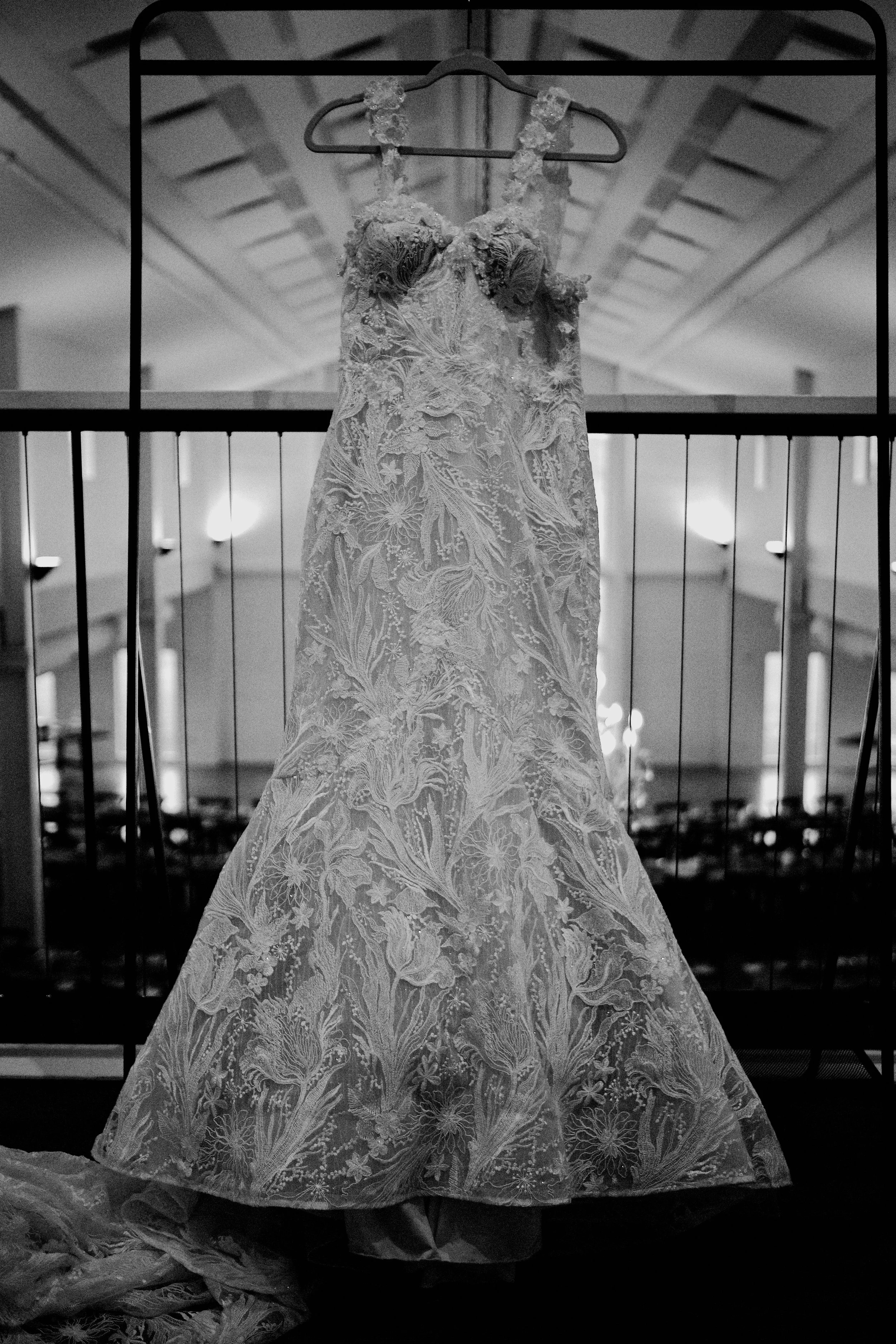 A wedding dress hanging on a hanger in an indoor space, with a decorative lace pattern, illuminated by soft lighting.