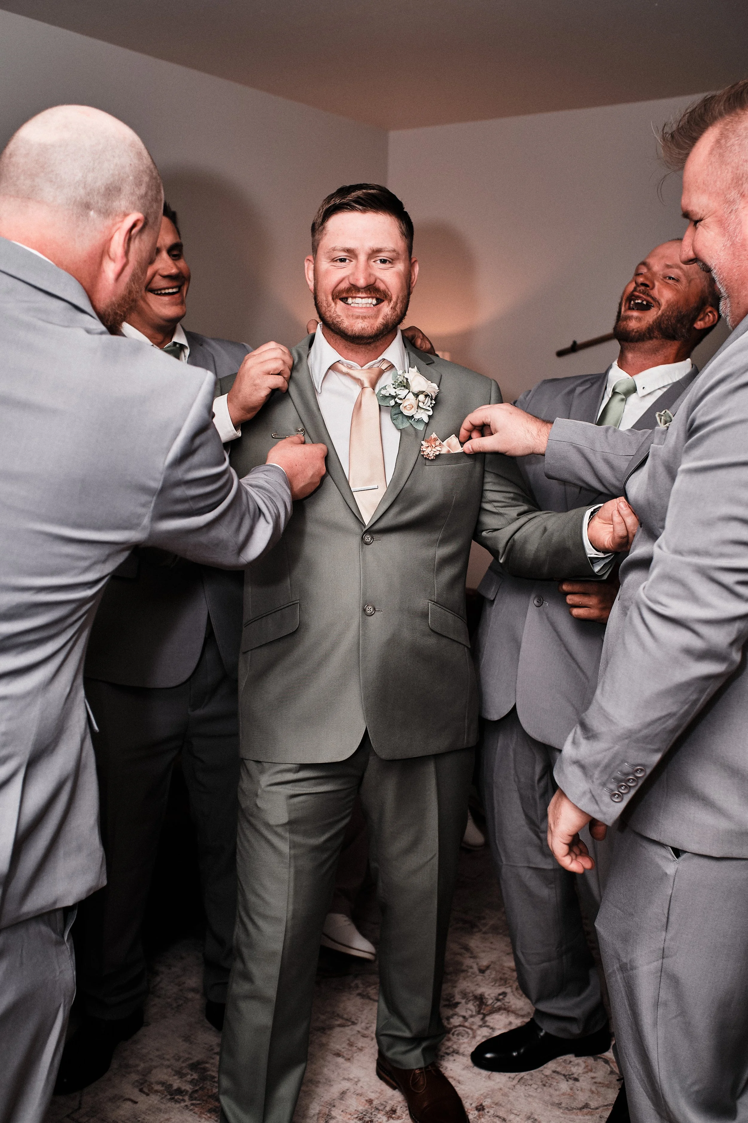 Groom in gray suit with boutonniere, smiling, surrounded by groomsmen adjusting his jacket at wedding celebration.