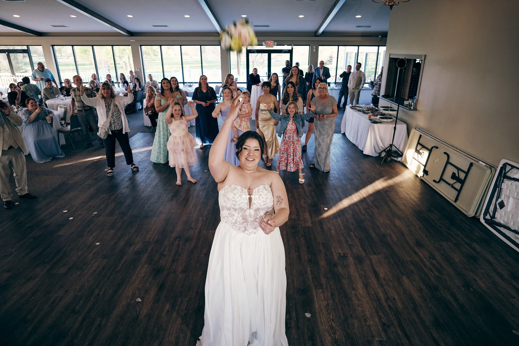A bride in a white wedding dress stands at the reception hall, smiling, with guests and family in the background, some raising their hands, and a bouquet above her head.