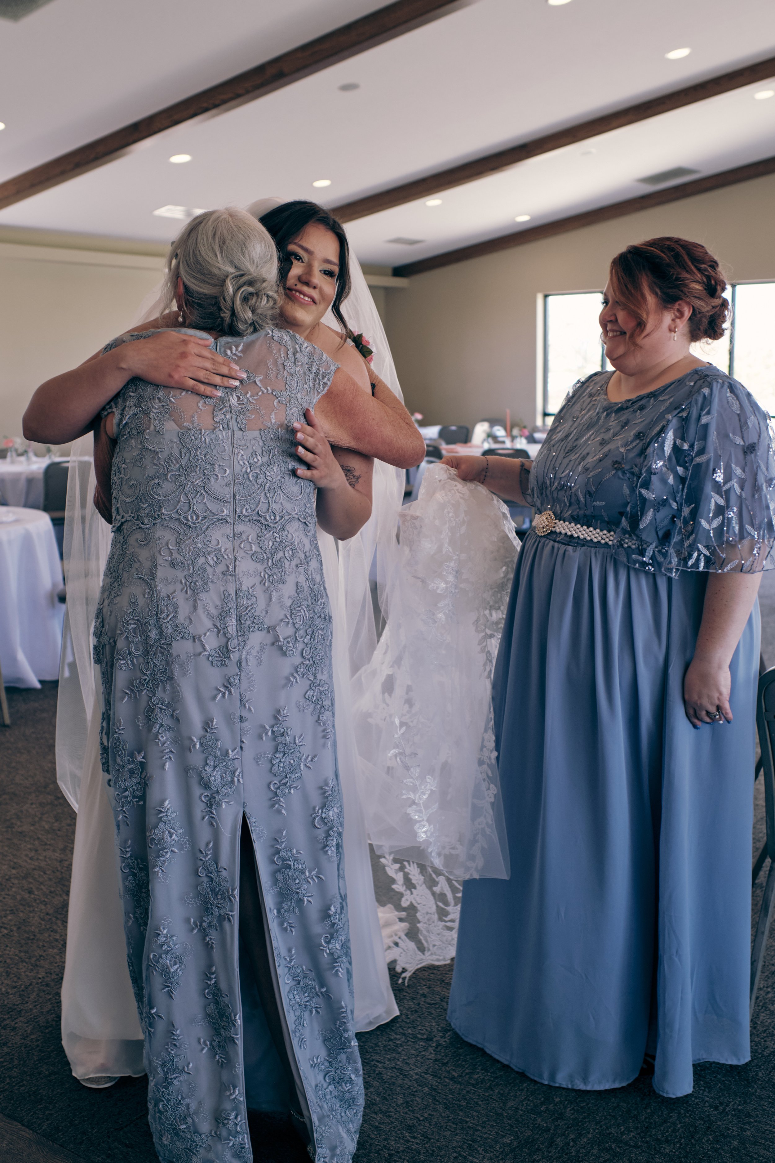 A bride in a white wedding dress is hugging an older woman in a lavender floral lace dress. Another woman in a blue lace dress is smiling and holding a lace wedding veil. The background shows a room with round tables and chairs, large windows, and ce