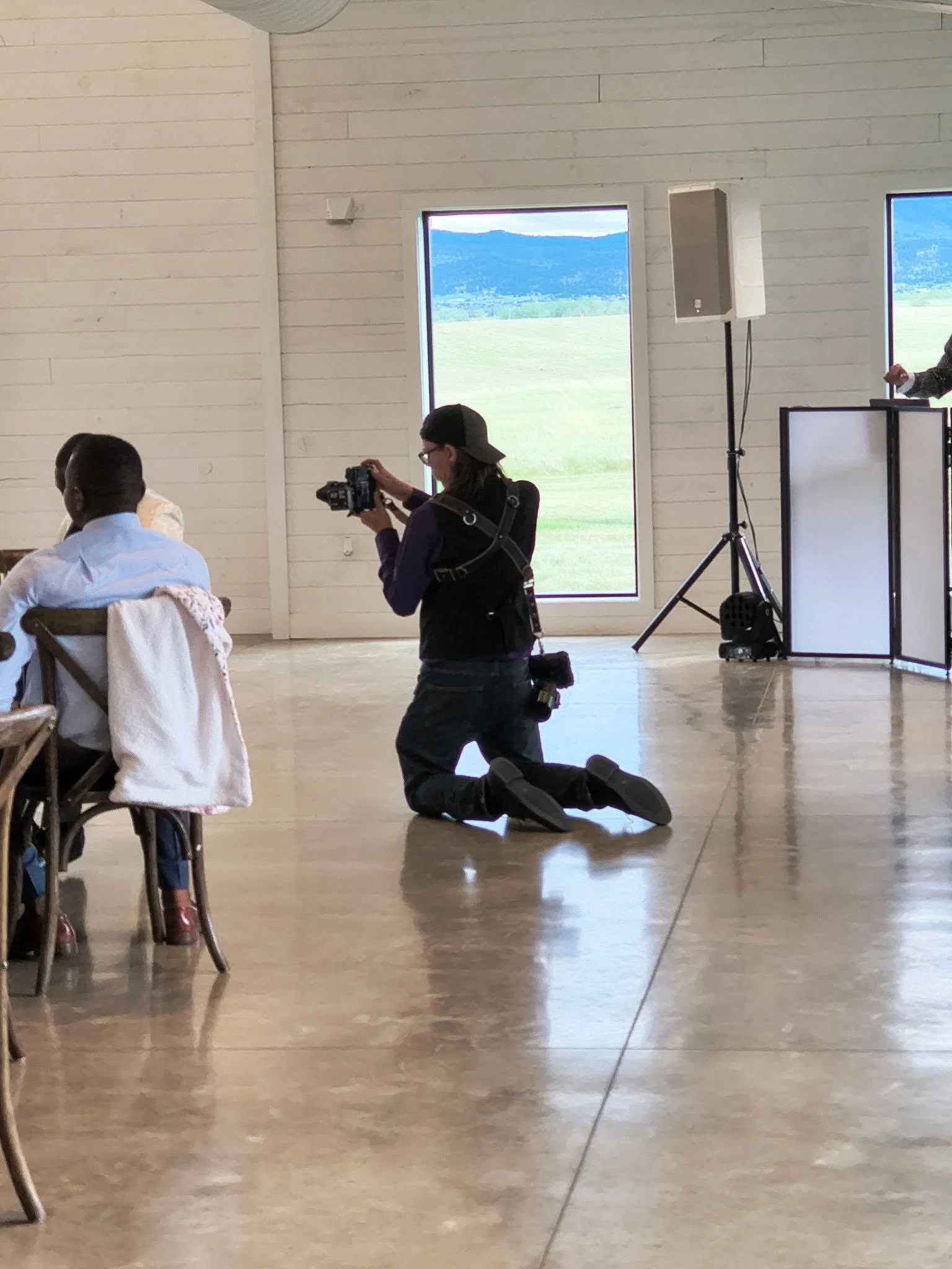 A photographer kneeling on the floor with a camera in hand in a large, well-lit room with white wooden walls and large windows showing a scenic landscape of mountains and fields outside. There are two people seated at a table on the left side of the image, and a speaker and a lighting stand are visible on the right.