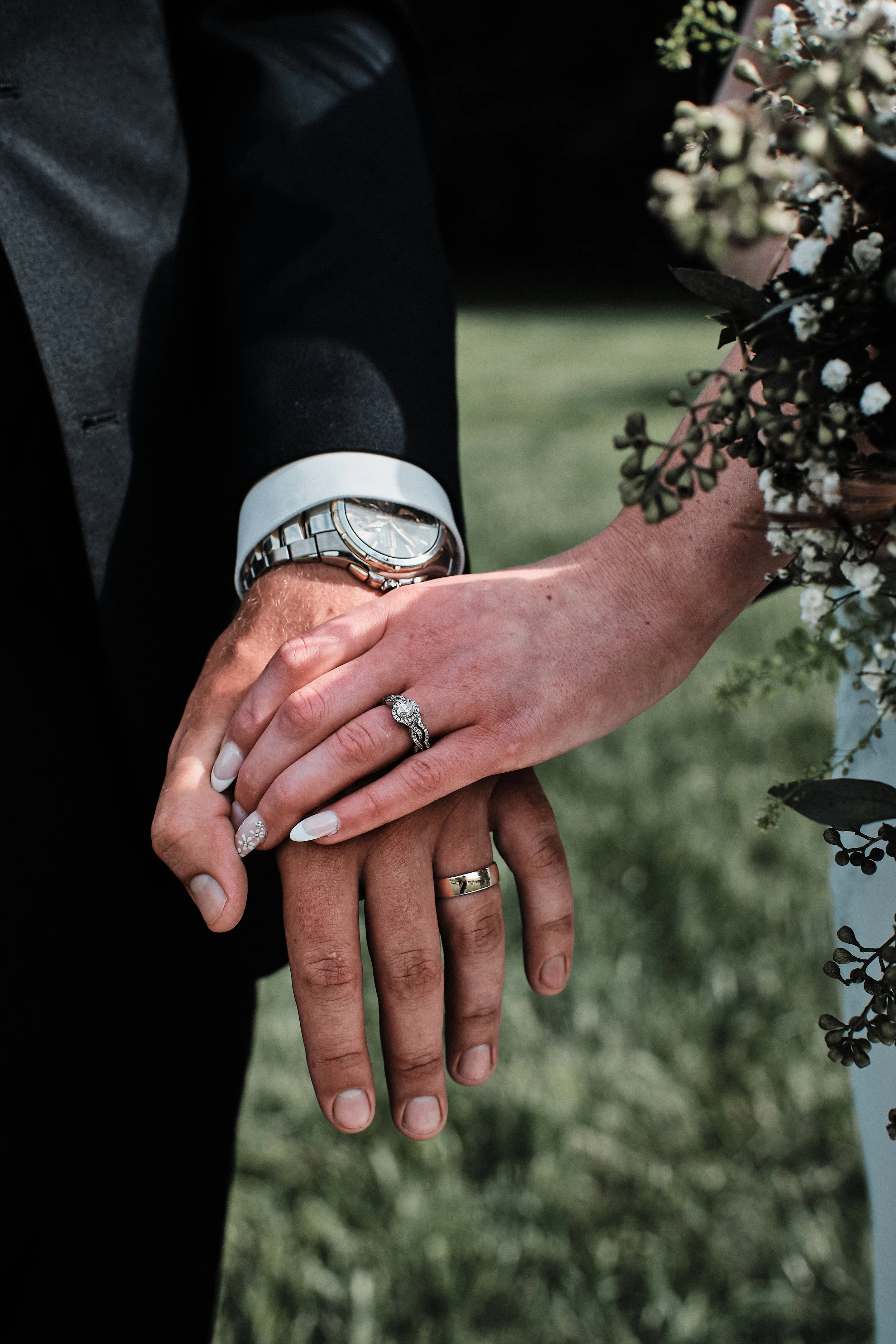 Close-up of two hands with wedding rings, one hand resting on top of the other. A wristwatch is visible on the person wearing a black suit, with a white shirt cuff showing. There are flowers in the background.