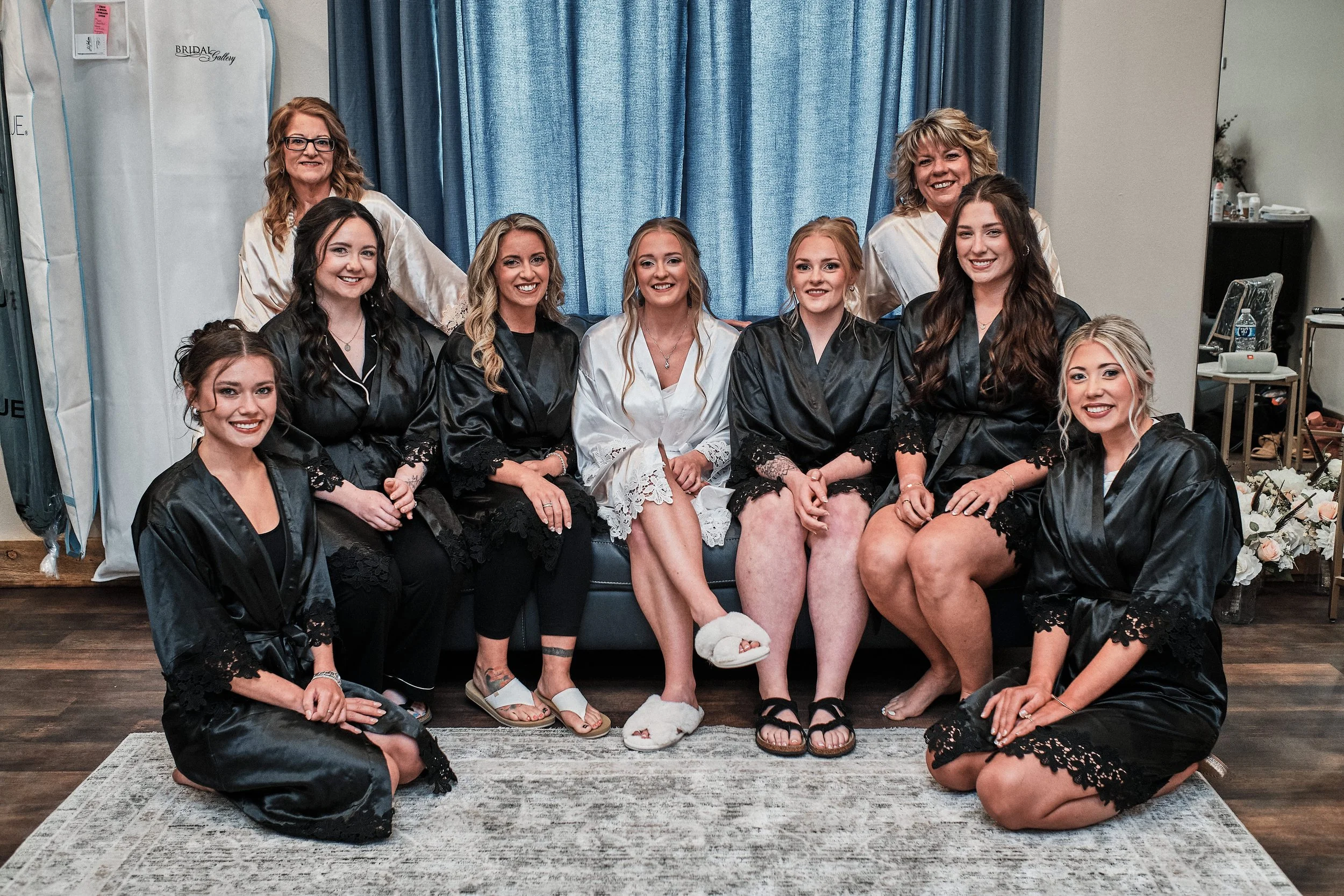 A group of nine women, some sitting and some standing, gathered in a room with blue curtains. They are wearing a mix of black and white robes, possibly for a wedding or special occasion, and are smiling at the camera. One woman in the middle is dress