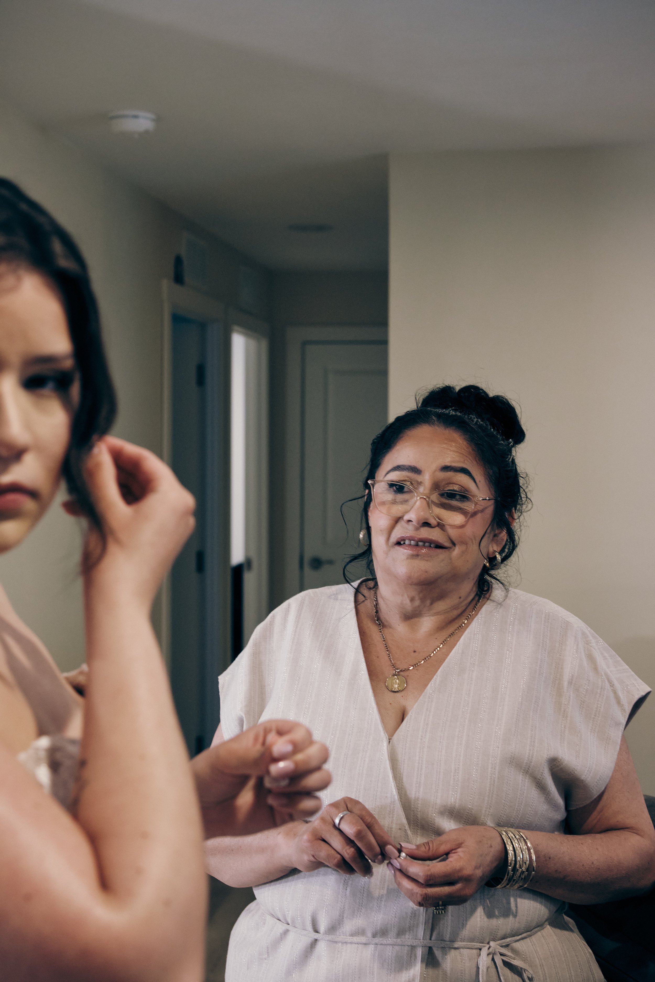 Two women are indoors, with one woman partially visible on the left side and the other woman in the center. The woman in the center has dark curly hair, wears glasses, a white short-sleeved top, and jewelry, and is smiling while talking or engaged wi