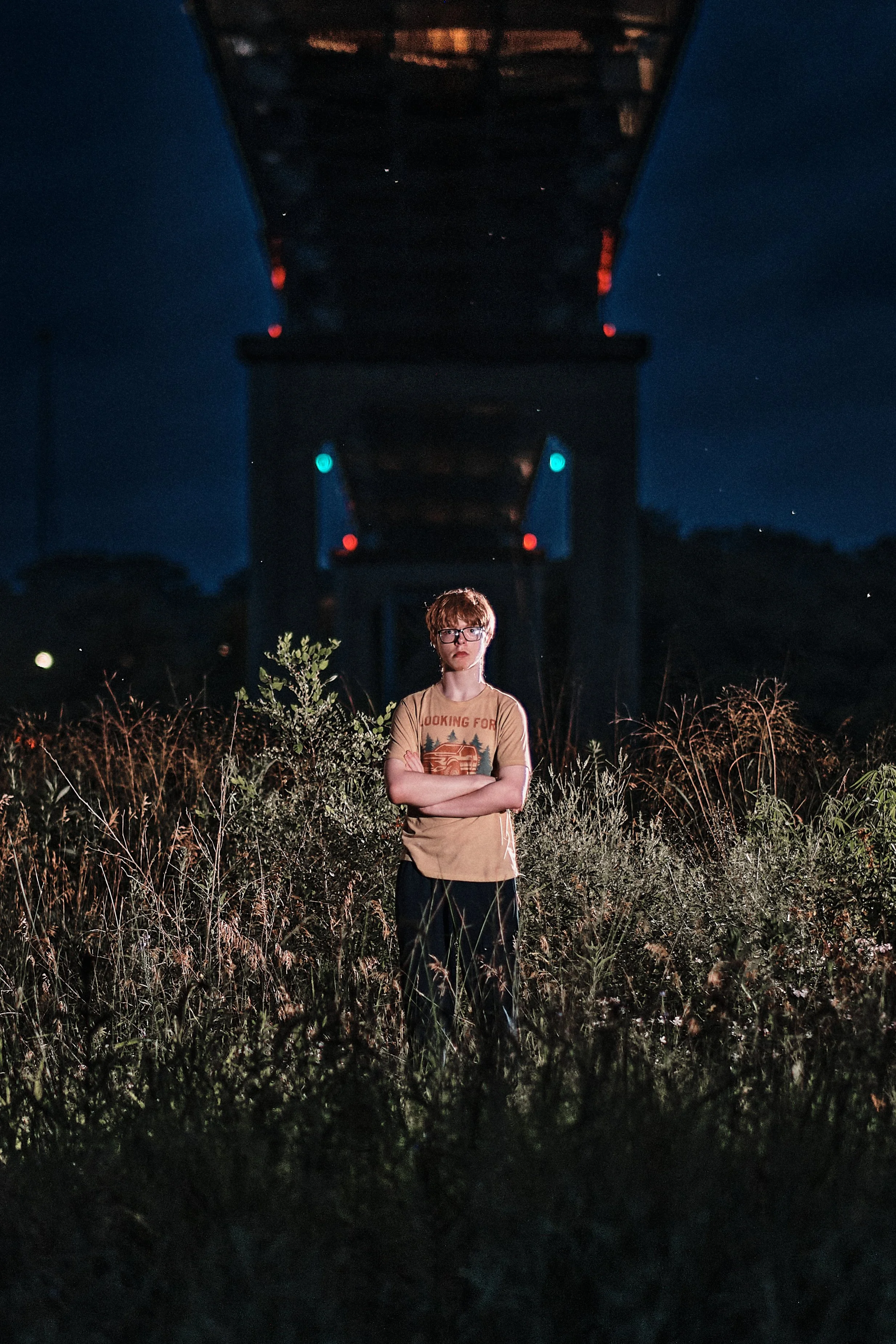 A young person with red hair and glasses standing with arms crossed in front of a dark bridge at night, surrounded by tall grass and bushes.