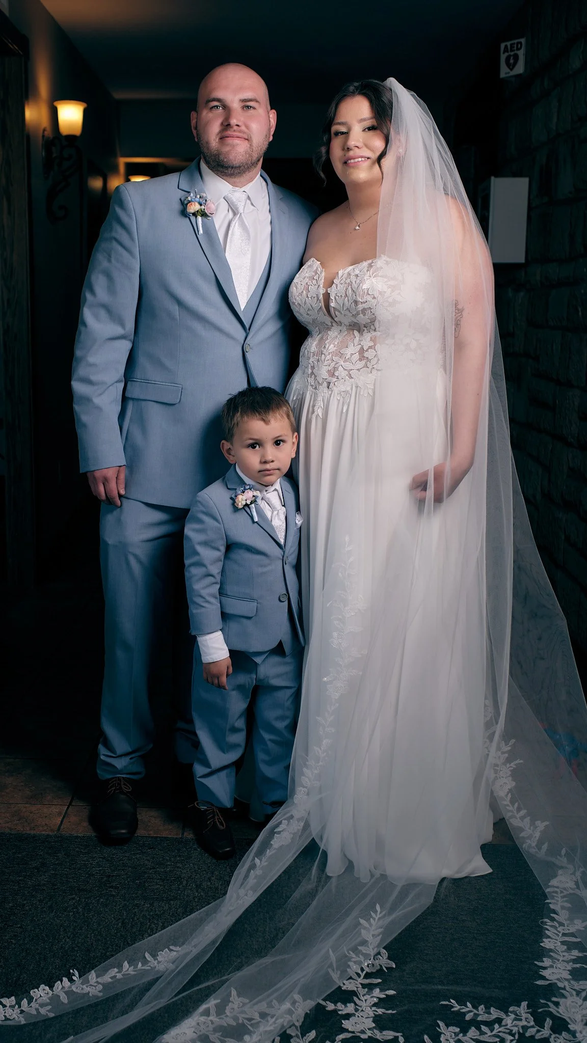 A bride in a white wedding dress and veil stands between a man and a young boy, all dressed in suits, in an indoor setting with dark walls and soft lighting.