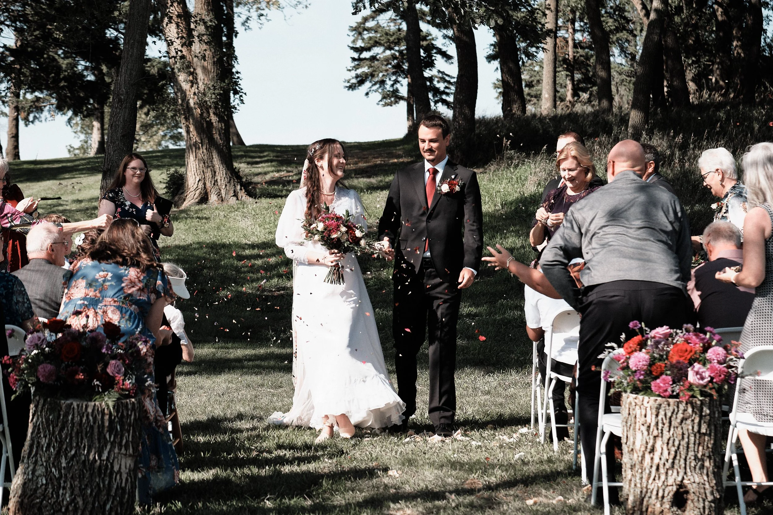 A bride and groom walking down the aisle at an outdoor wedding ceremony with guests seated on either side, trees in the background, flower arrangements, and petals on the grass.