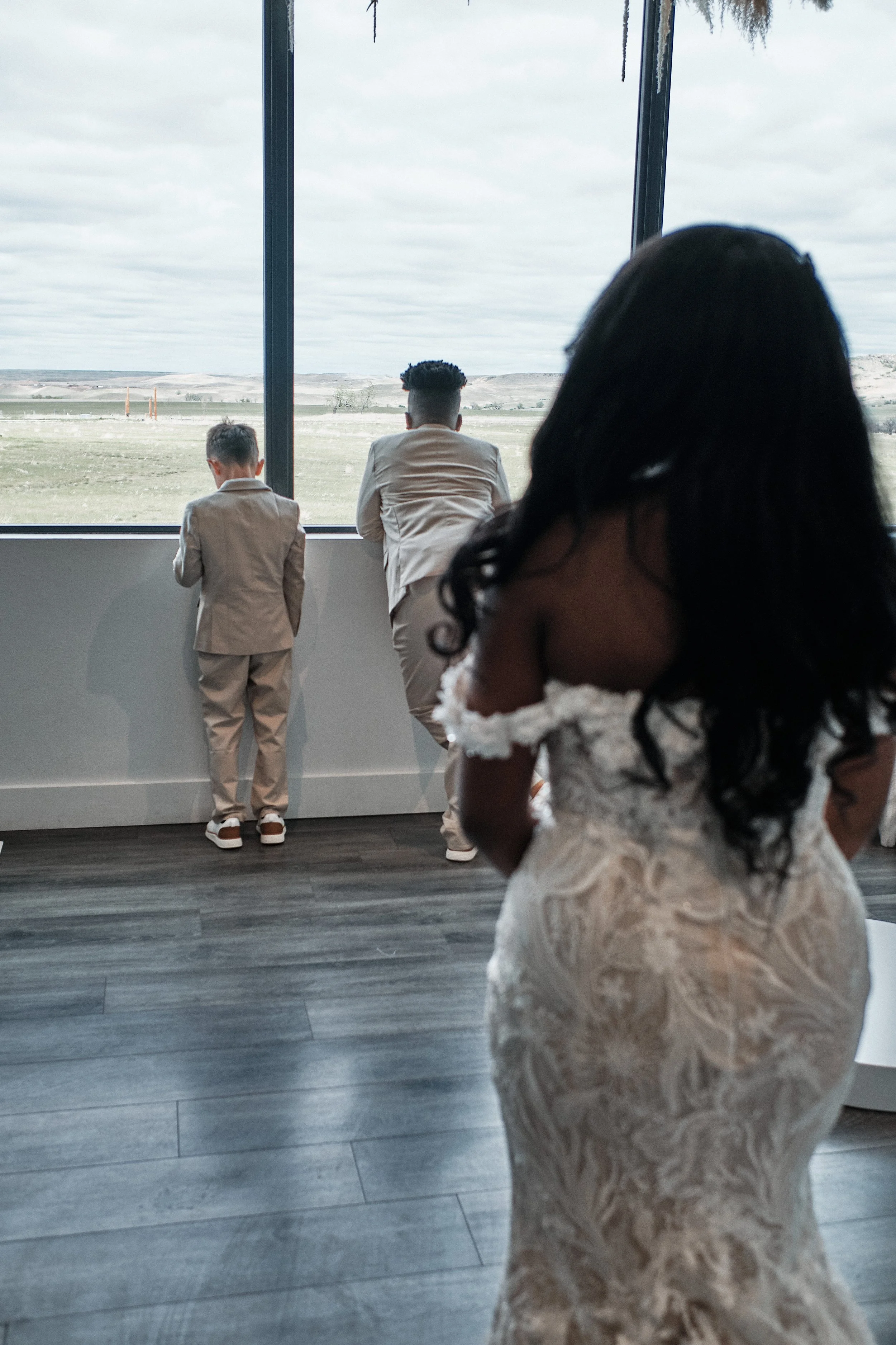 A group of people, including two boys in tan suits and a woman in a white lace dress, are standing indoors and looking out through a large window at an open landscape with fields and a cloudy sky.