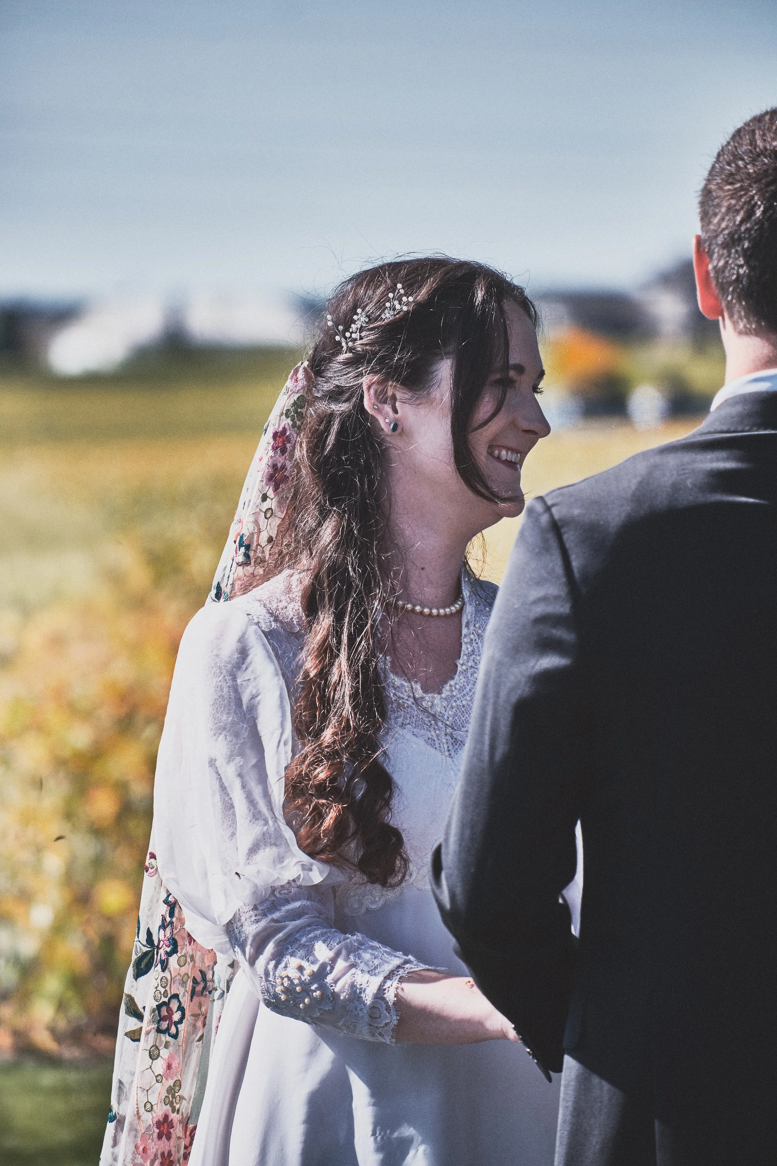 A bride with long, dark, curly hair wearing a floral embroidered veil and a white lace wedding dress, smiling while holding hands with a groom in a dark suit outdoors.
