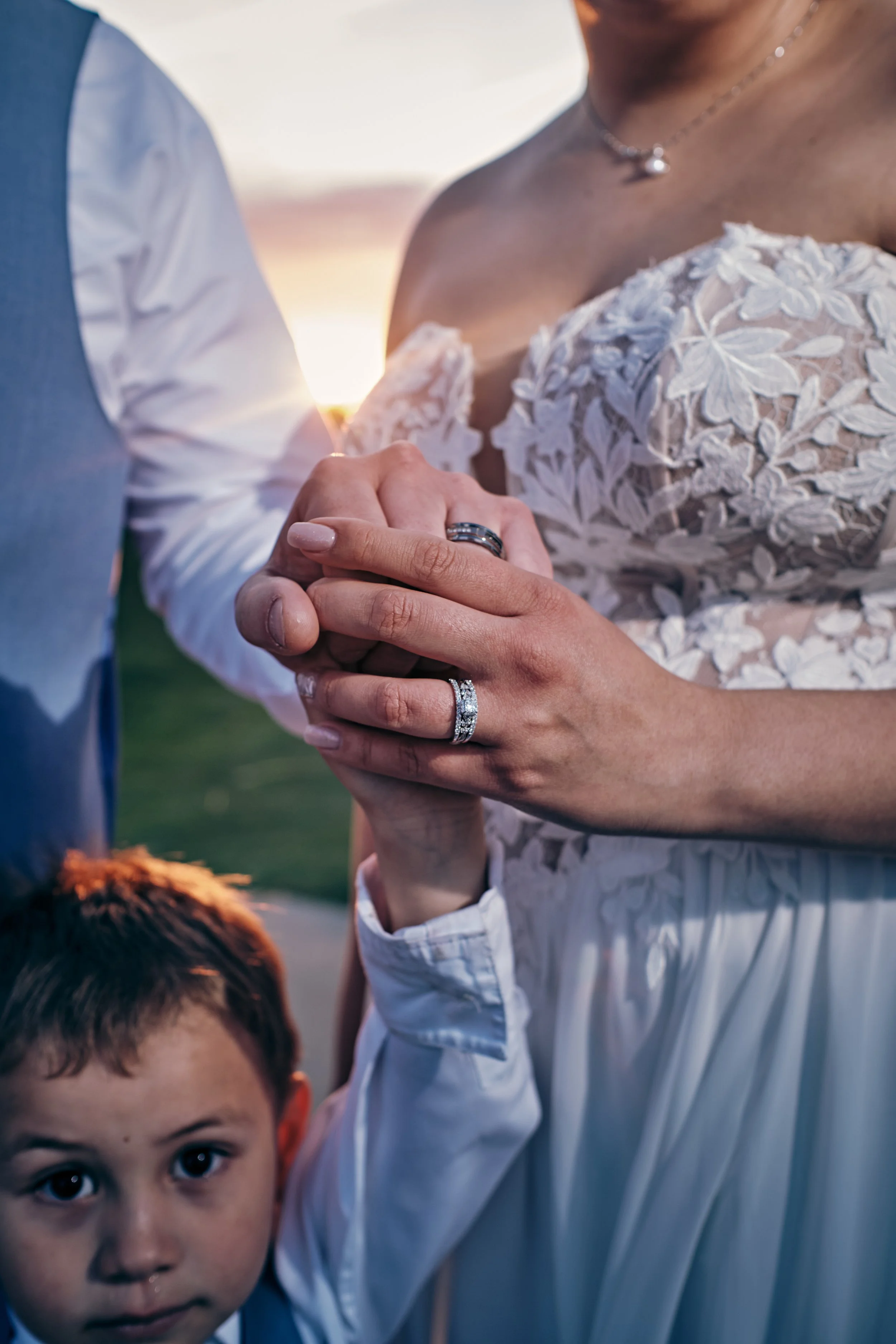 A bride and groom holding hands with wedding rings during their wedding ceremony, with a young child looking directly at the camera in the foreground.