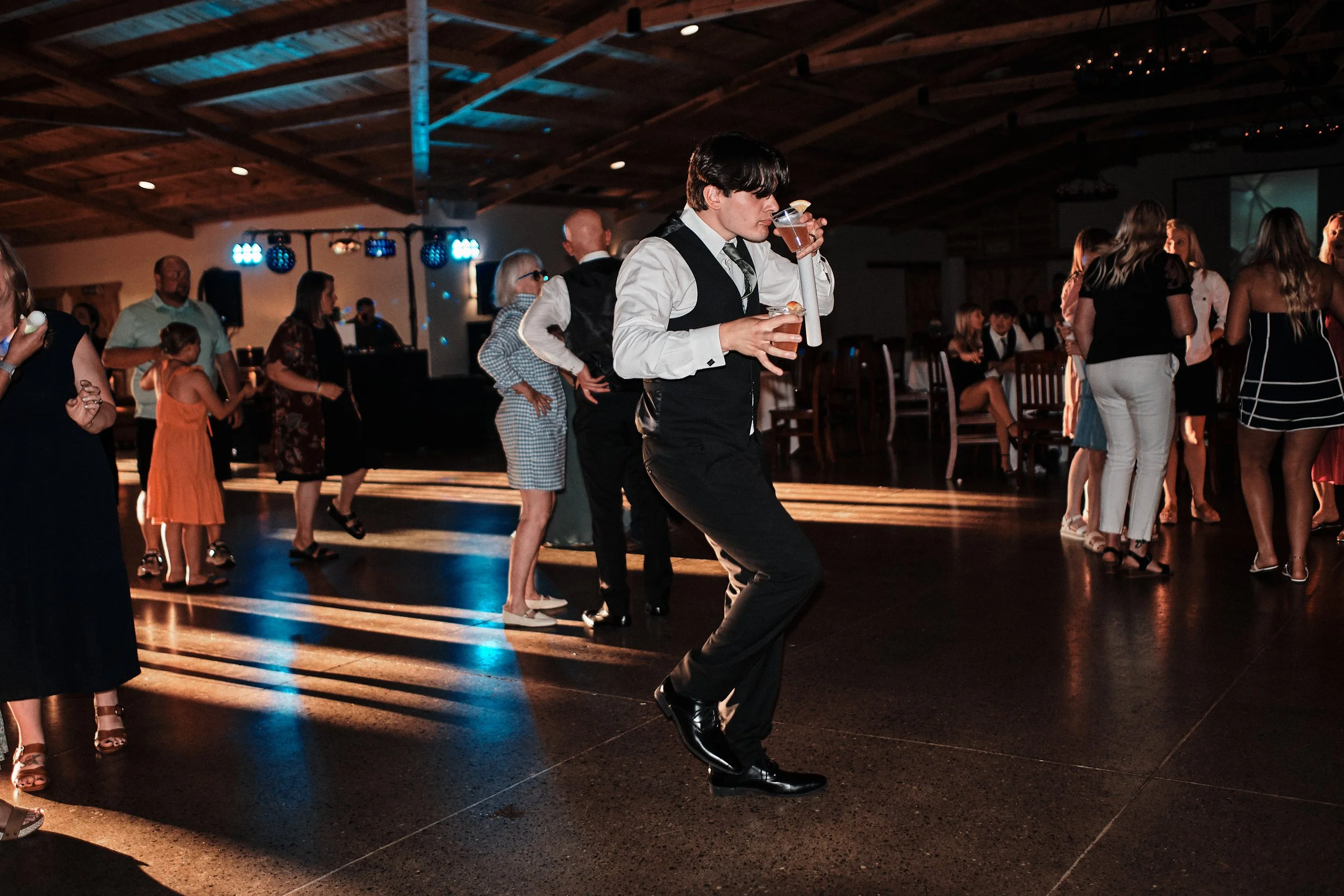 Young man in formal attire dancing at a social event, holding a drink, in a dimly lit venue with other people dancing and socializing in the background.