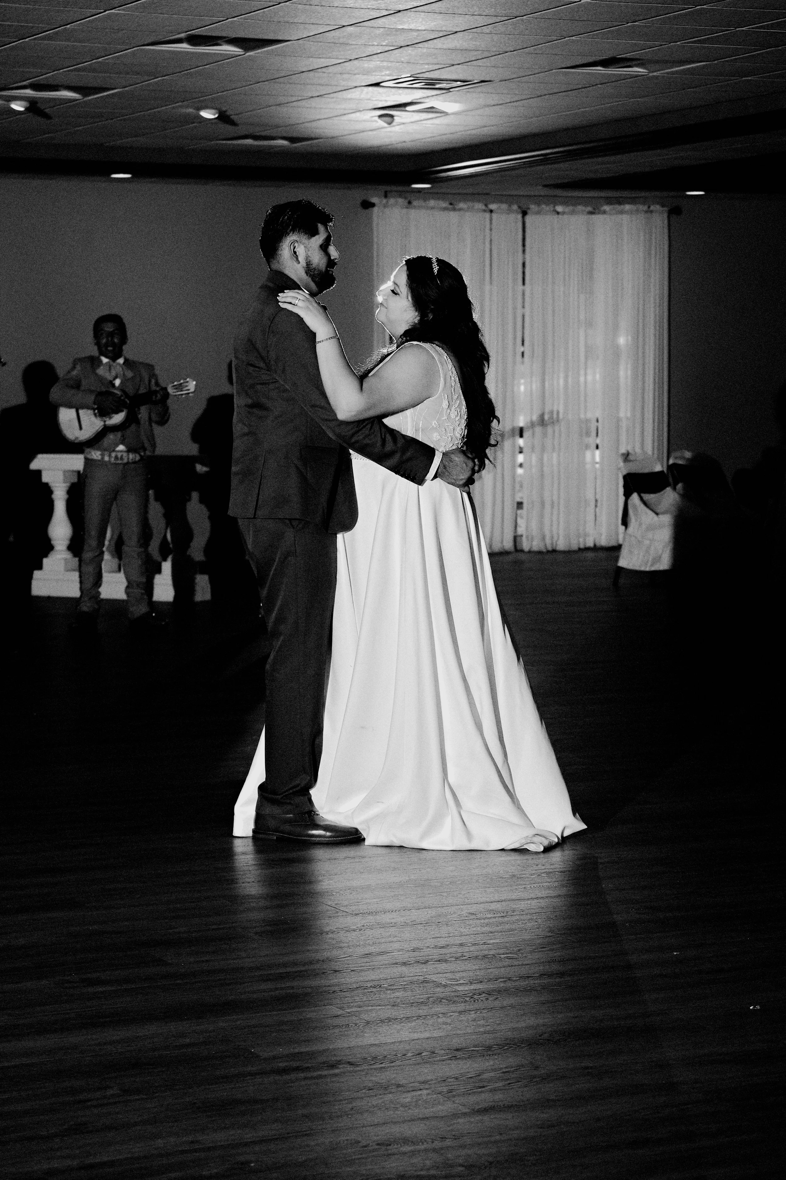 A couple dancing closely at an indoor wedding reception, with a musician playing guitar in the background and guests seated at tables.
