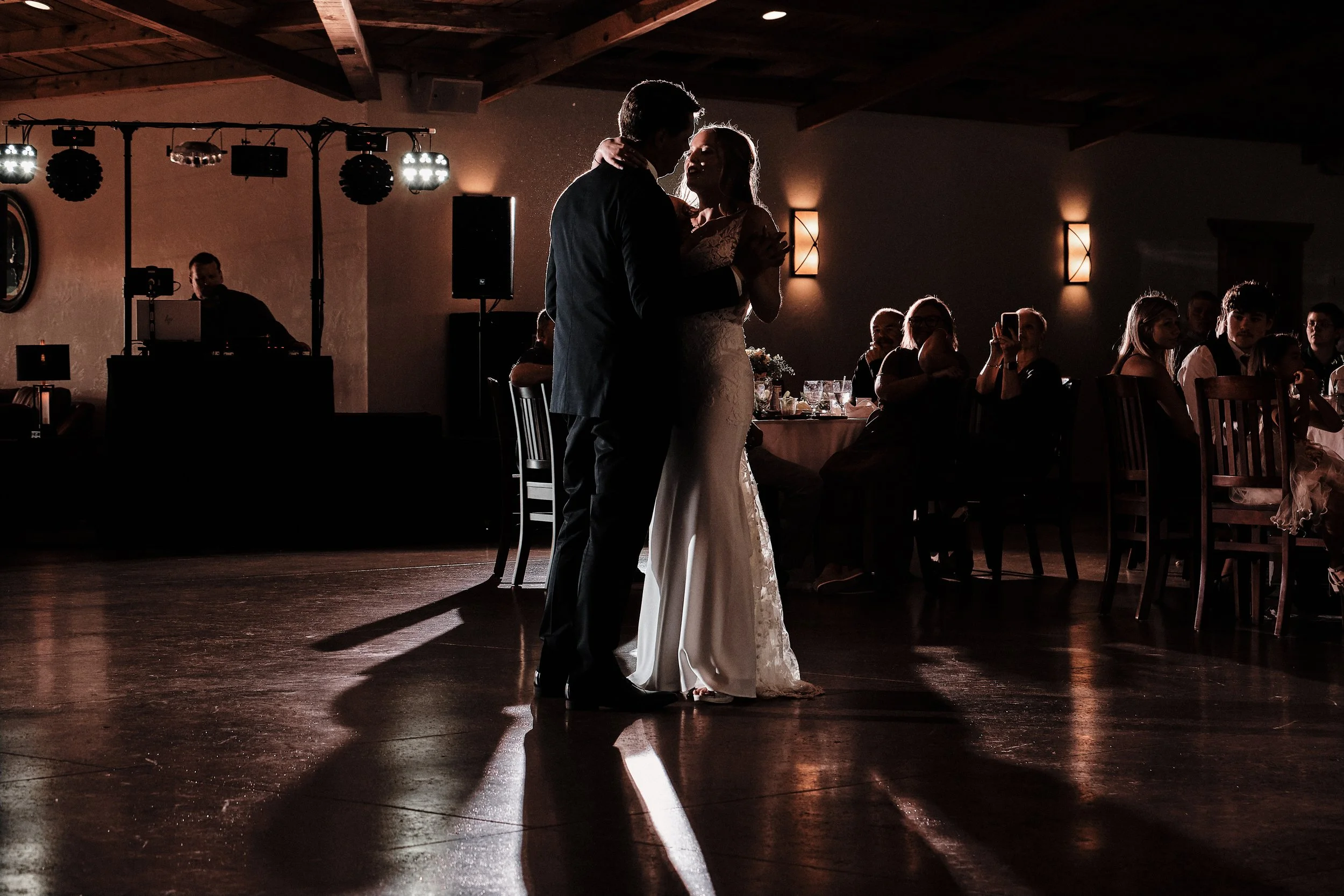 A newlywed couple sharing a dance at their wedding reception, illuminated by warm lighting with guests watching.