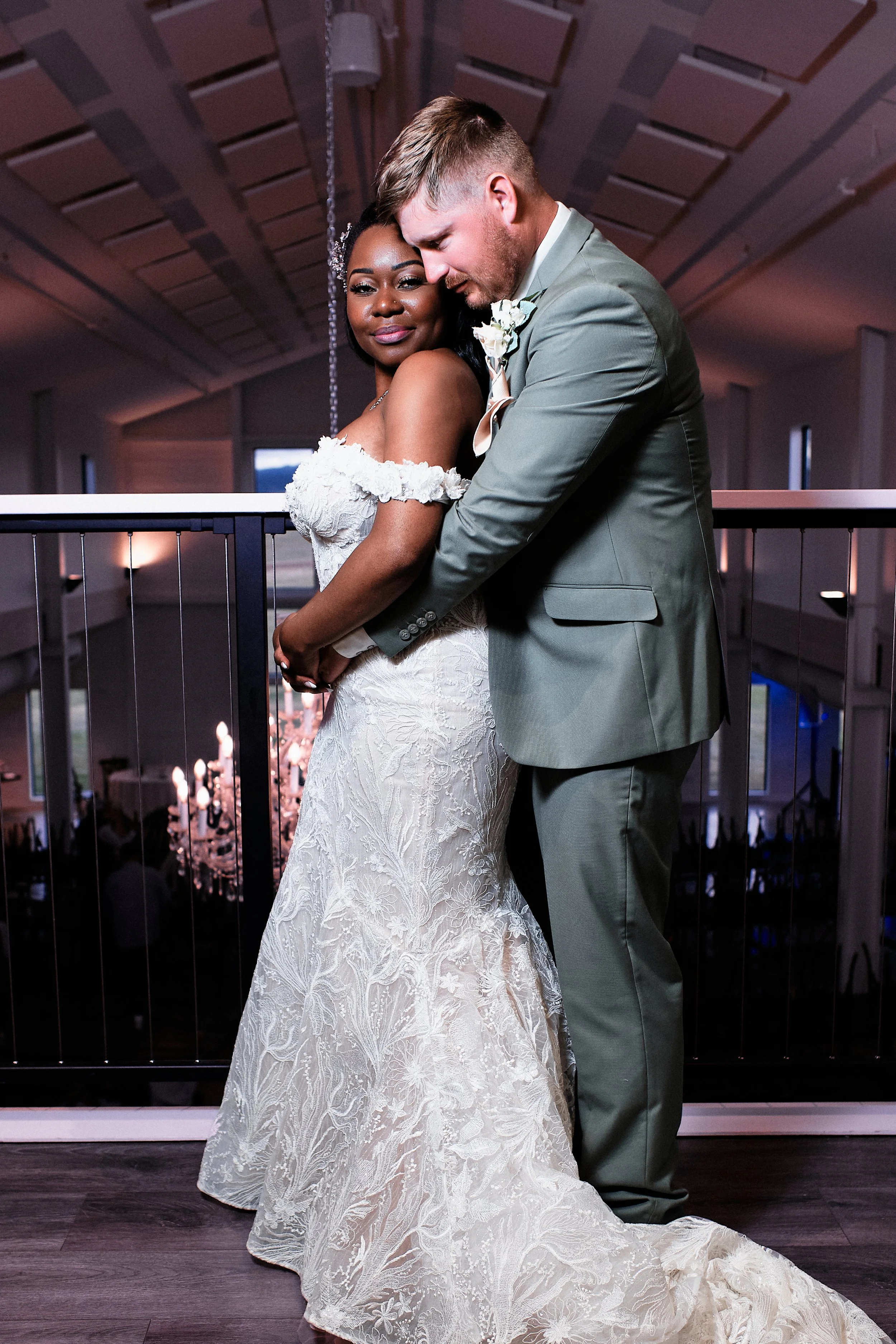 A bride in a white lace wedding gown and a groom in a gray suit dancing closely at their wedding reception, with a chandelier and guest tables visible in the background.