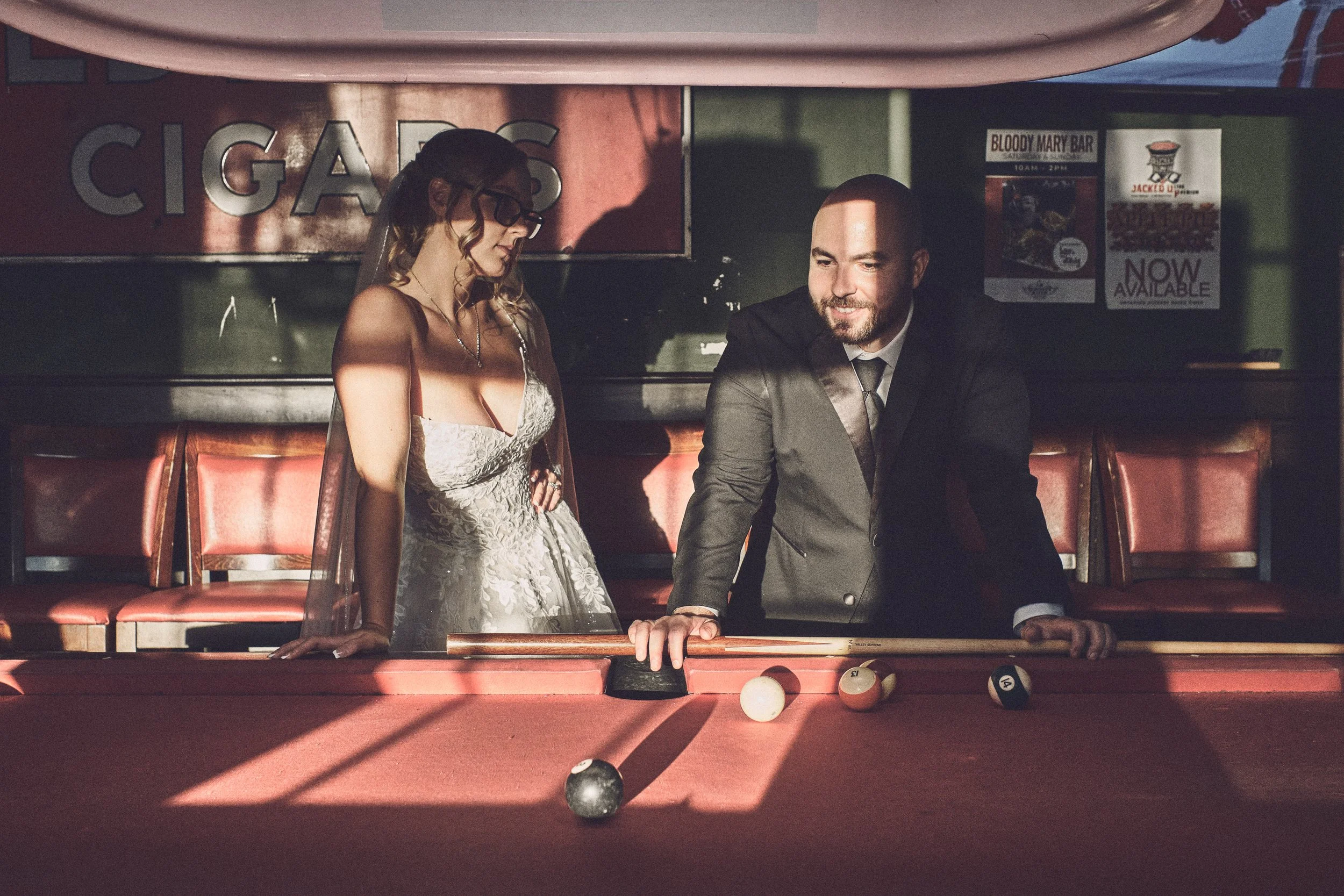 A woman in a wedding dress and a man in a suit playing pool at a billiards table in a bar or recreational area.