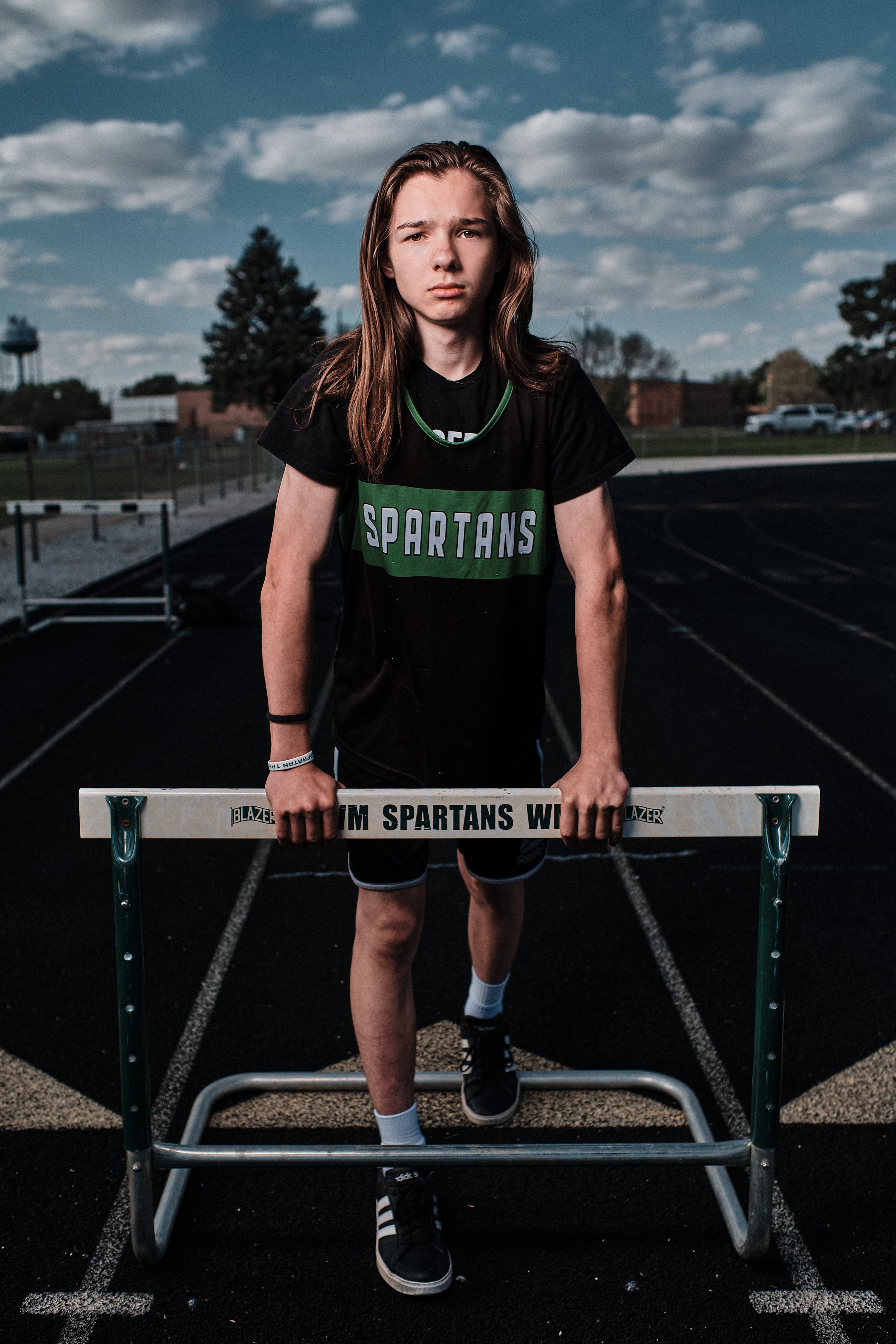 A young male athlete standing at a track starting block, wearing a black and green sports uniform with 'Spartans' written on it, at an outdoor athletic track during daytime with a cloudy sky.