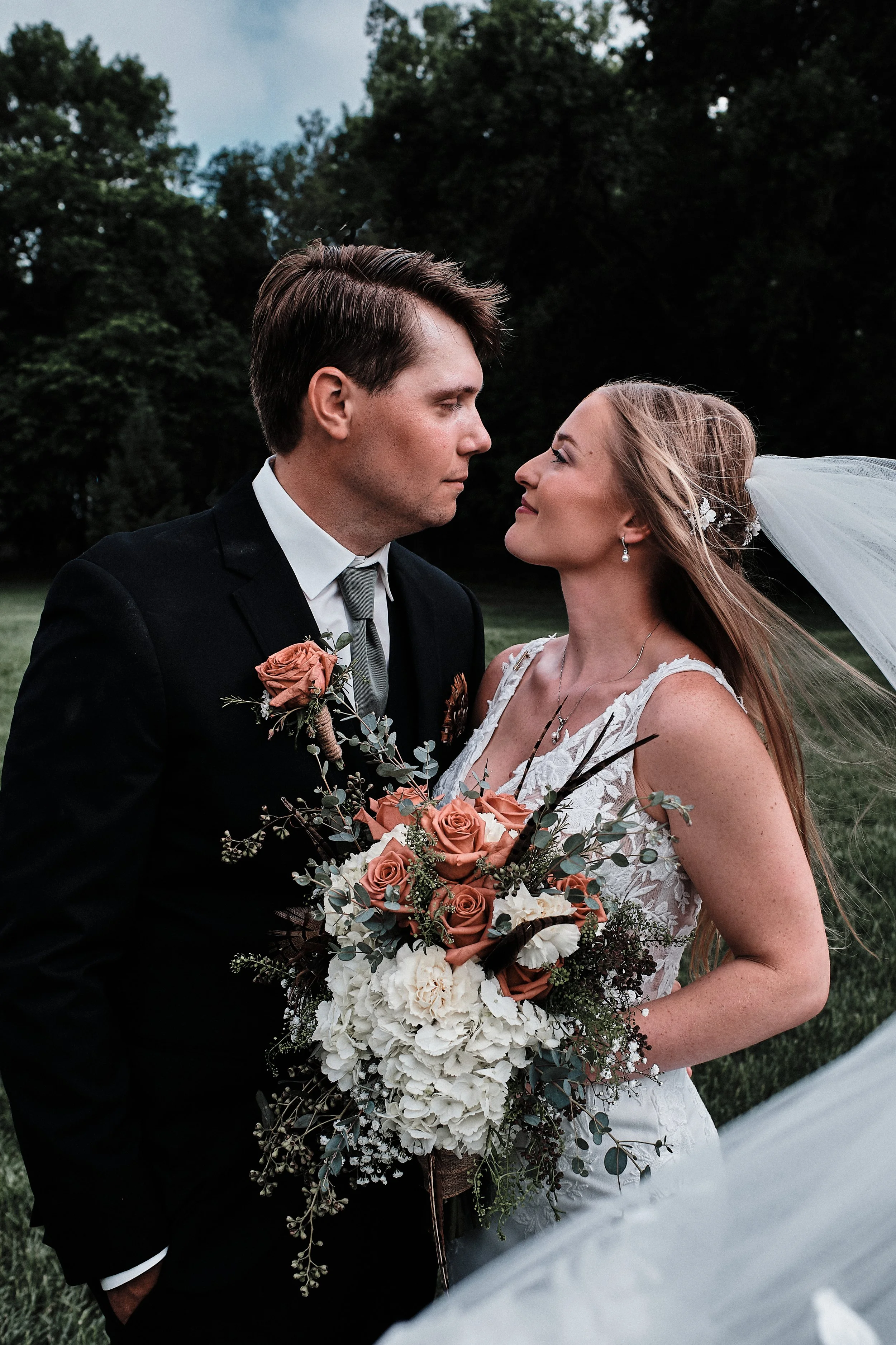 A bride and groom face each other outdoors during their wedding ceremony. The groom is dressed in a black suit with a gray tie, holding a bouquet of peach and white flowers. The bride wears a white lace wedding dress with a veil and earrings, holding