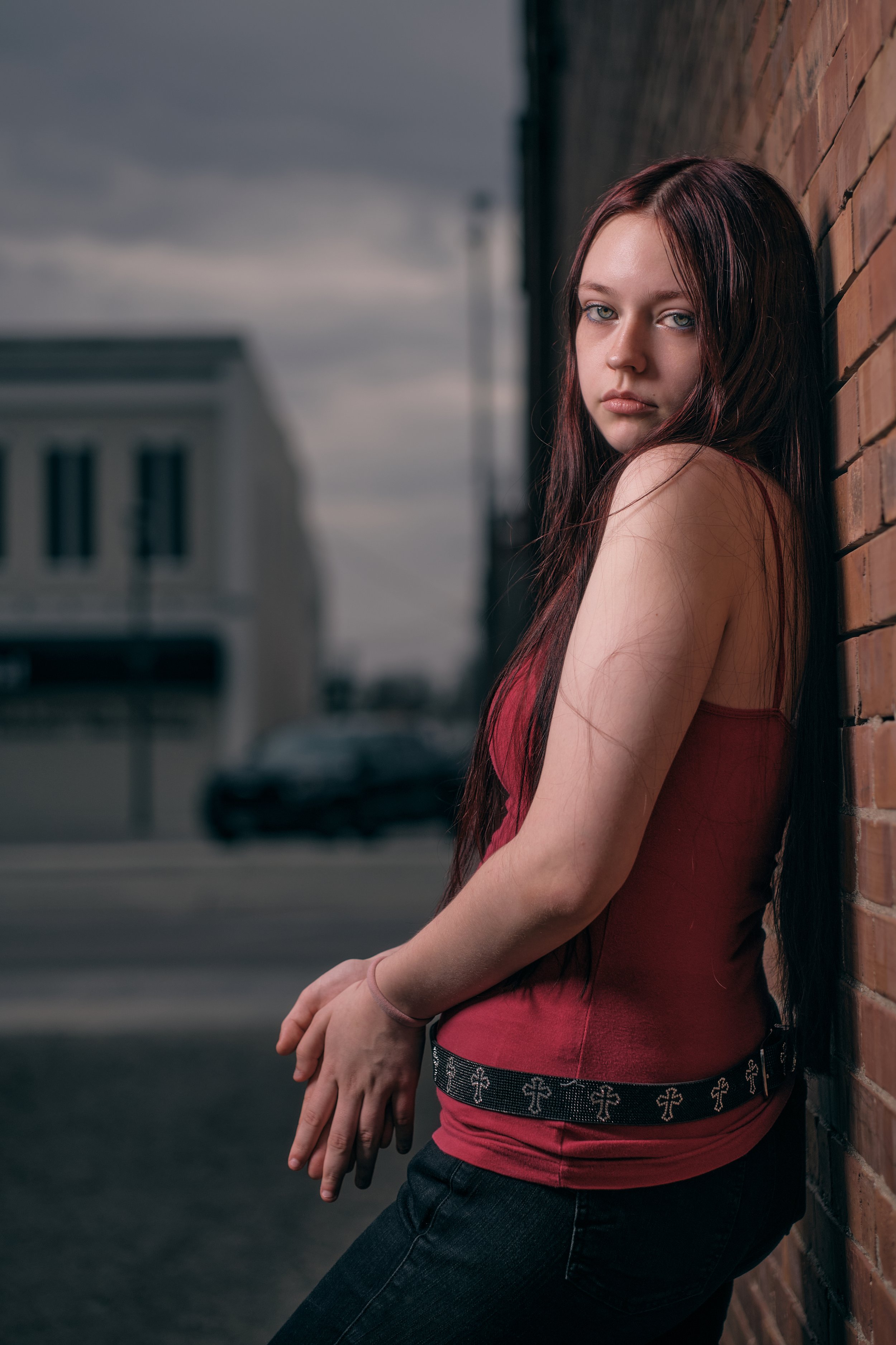 Young woman with long dark red hair and blue eyes leaning against a brick wall outdoors, wearing a red tank top and dark jeans, with a serious expression.
