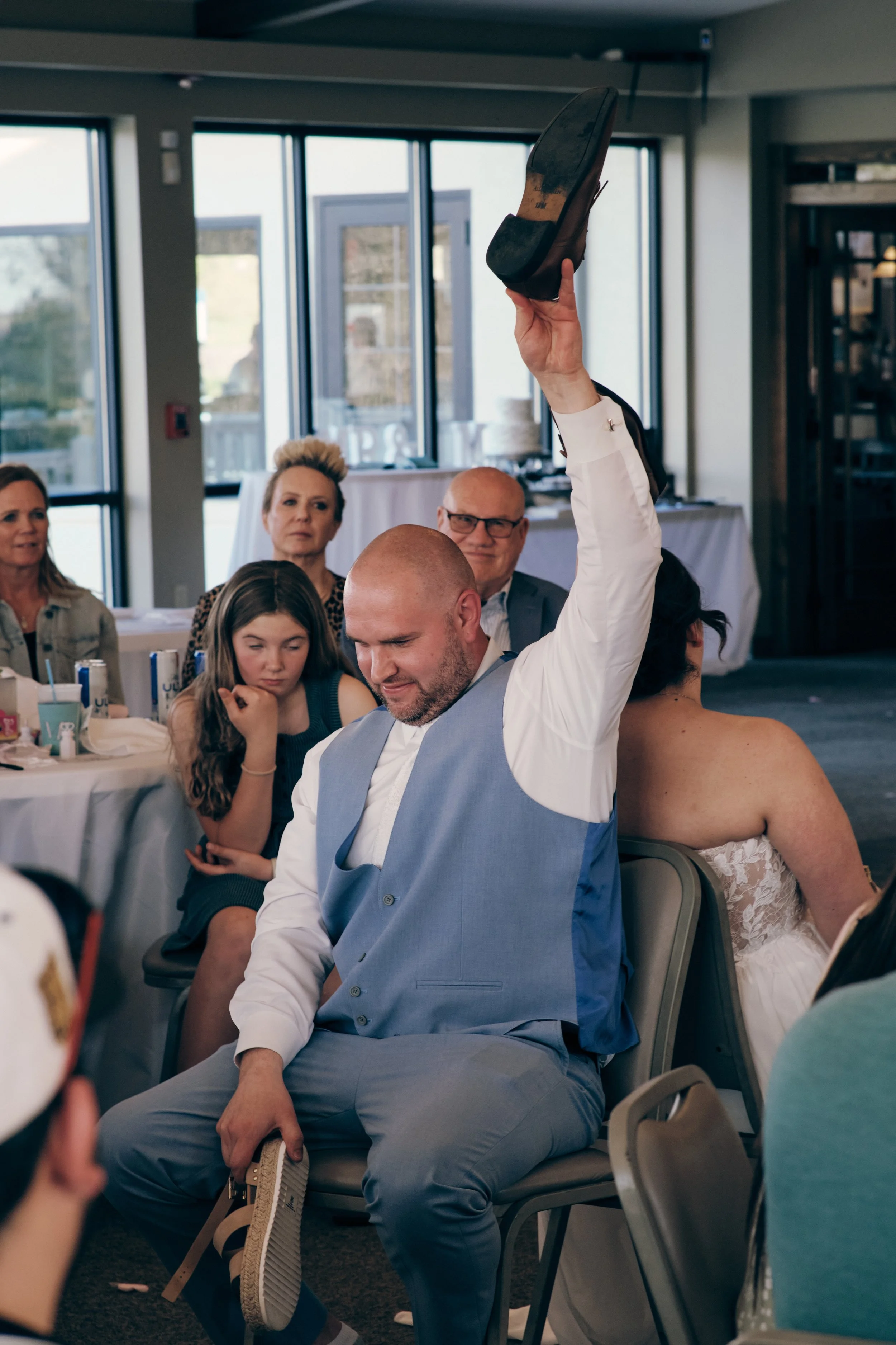 A man raising his shoe in a gesture during a gathering at a banquet hall with several seated guests observing him.