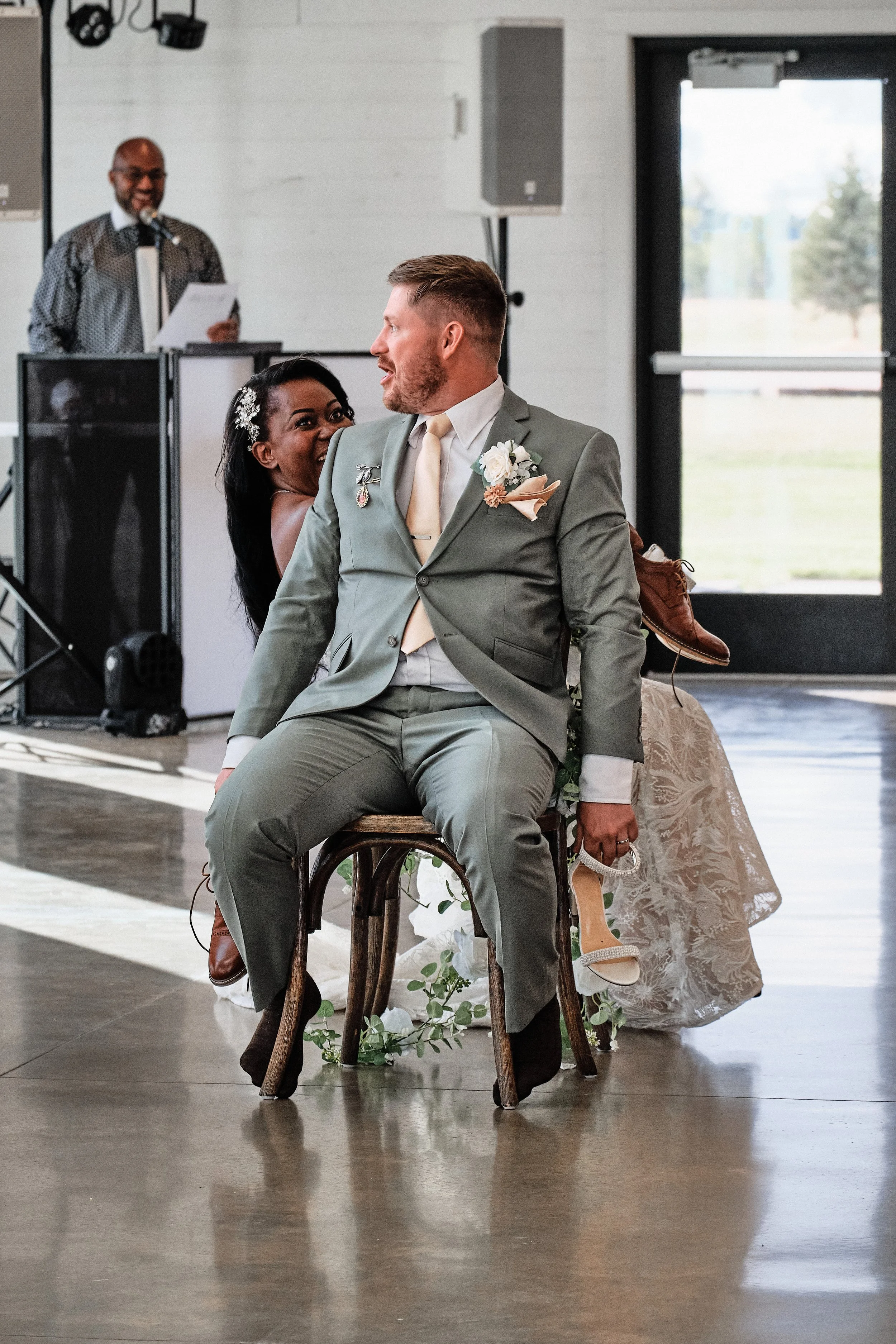 A newlywed couple dancing at their wedding reception, with the bride smiling and the groom looking at her, in a venue with natural light coming through large doors. A DJ is seen in the background.