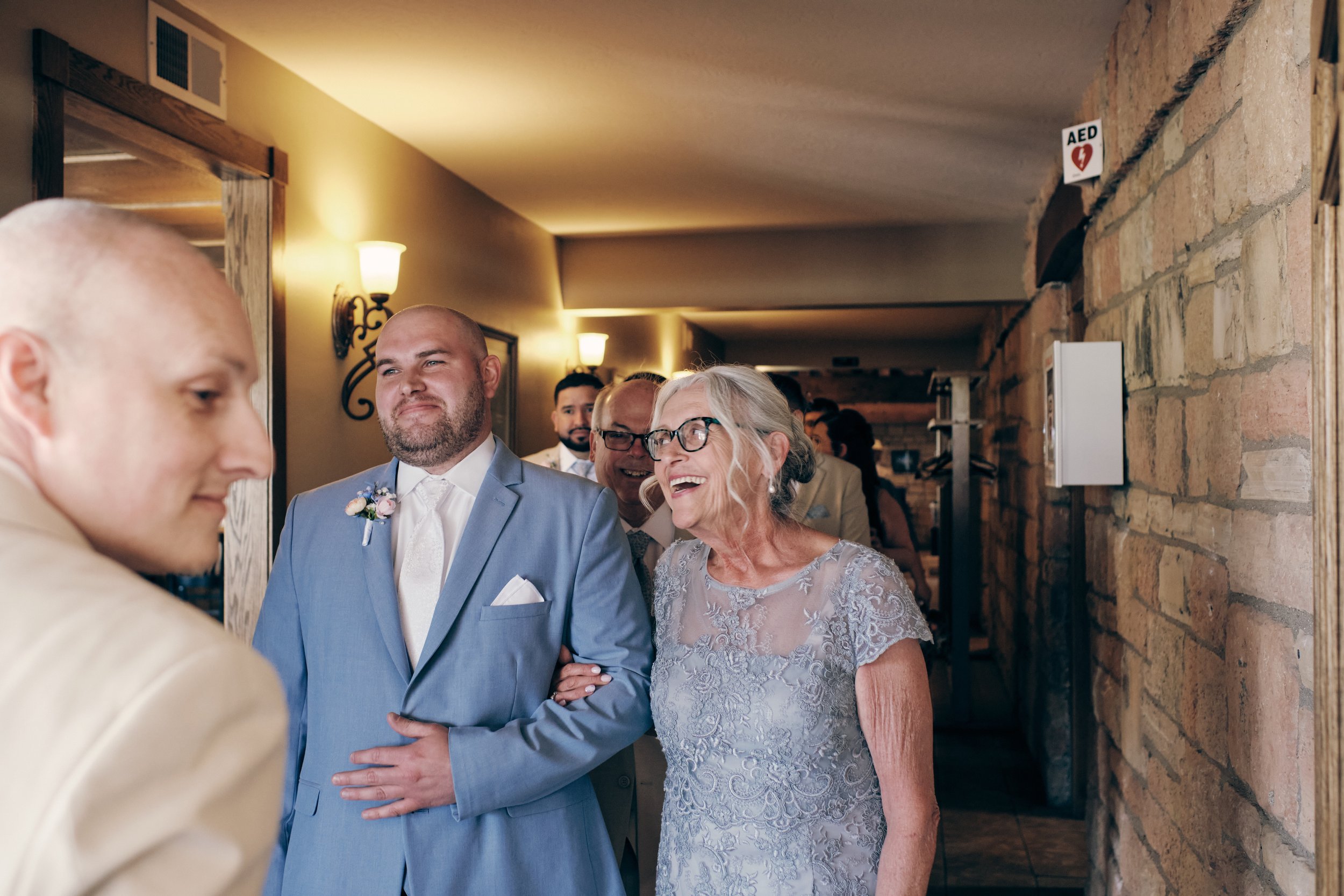 A group of people smiling and talking at a wedding reception. The groom is wearing a light blue suit, and an elderly woman in a lace dress is holding his arm. The setting is a warmly lit indoor venue with brick walls and wall-mounted lamps. An AED si