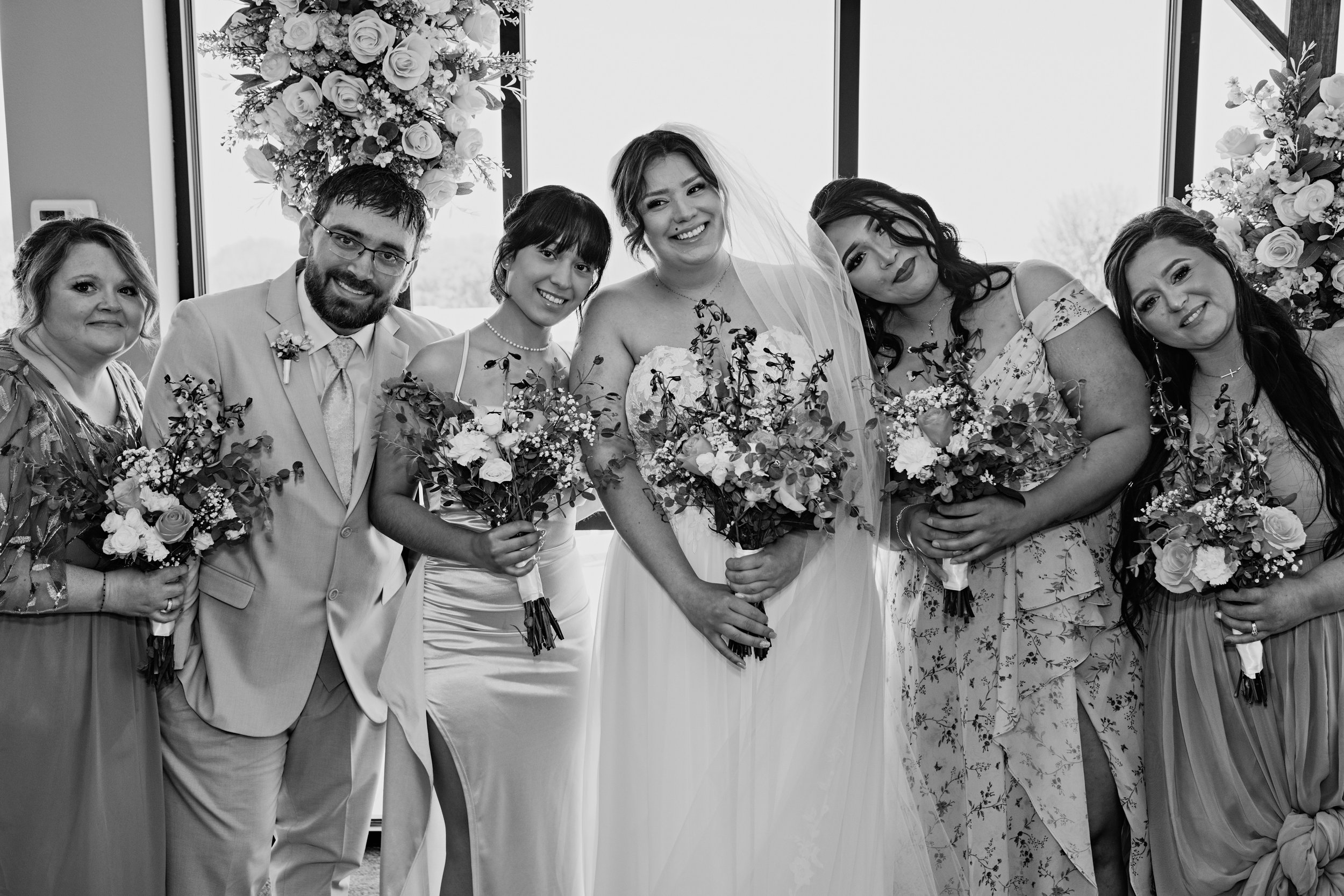 Black and white photo of six women and one man standing indoors, smiling, with the bride in the center. The women are holding bouquets of flowers, and the background features large windows with floral arrangements.