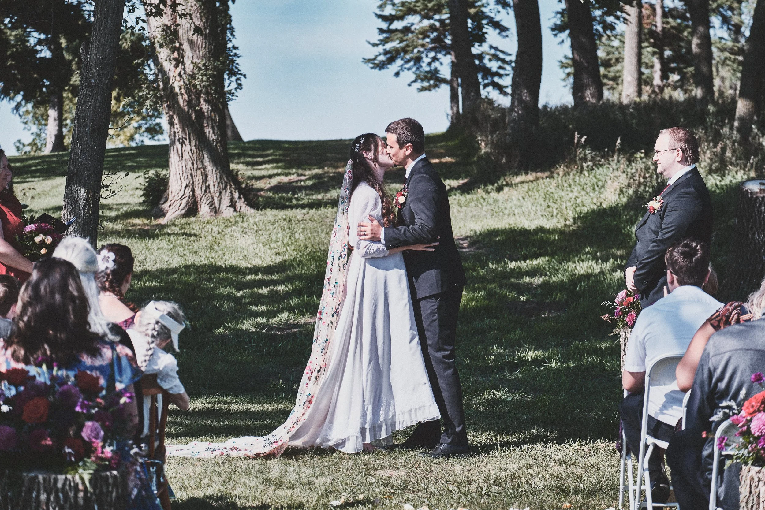 A wedding ceremony outdoors in a forested area with trees. The bride and groom are kissing, surrounded by seated guests dressed in colorful clothing.