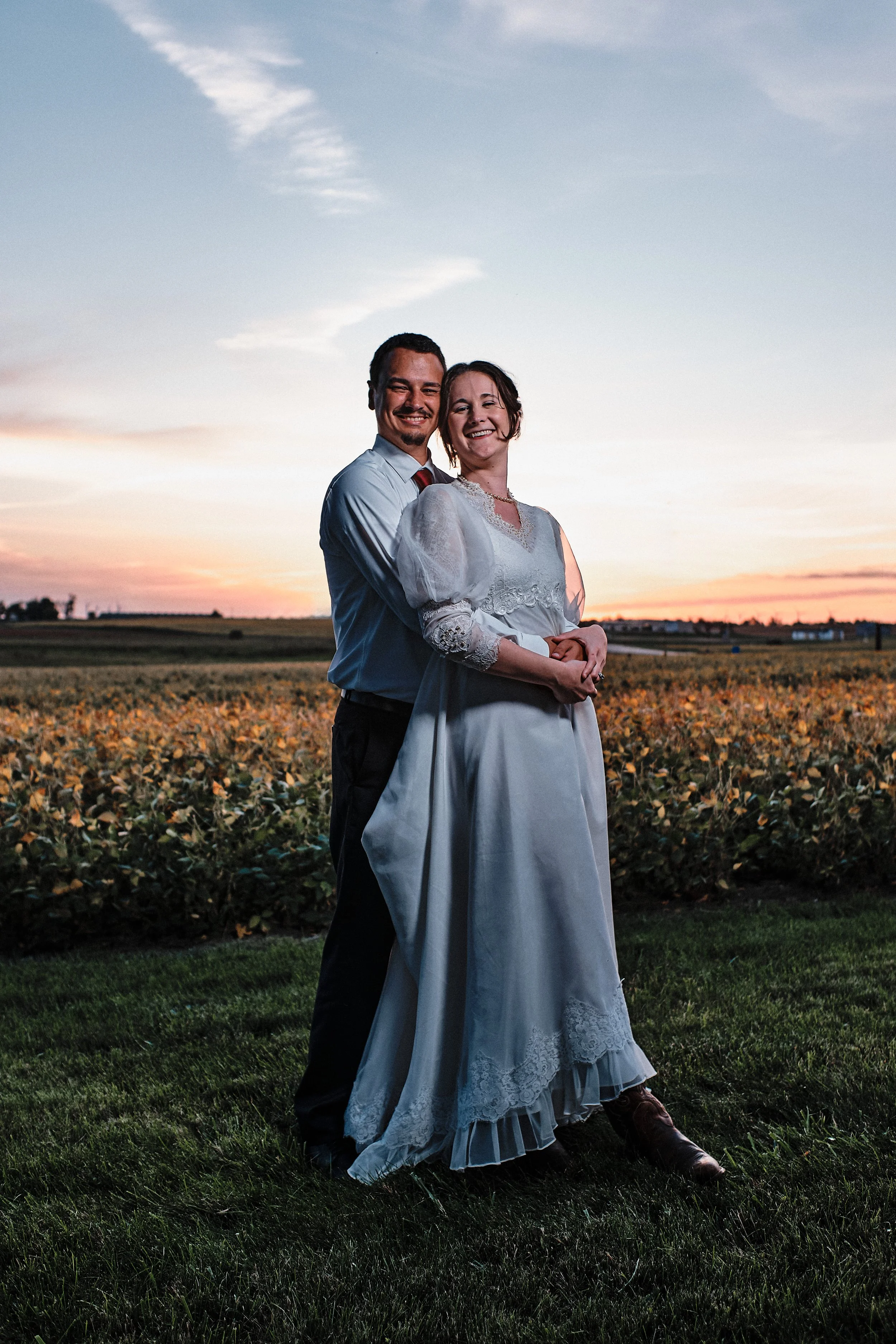 A smiling man and woman in wedding attire standing together in a field at sunset, with a clear sky and distant trees.