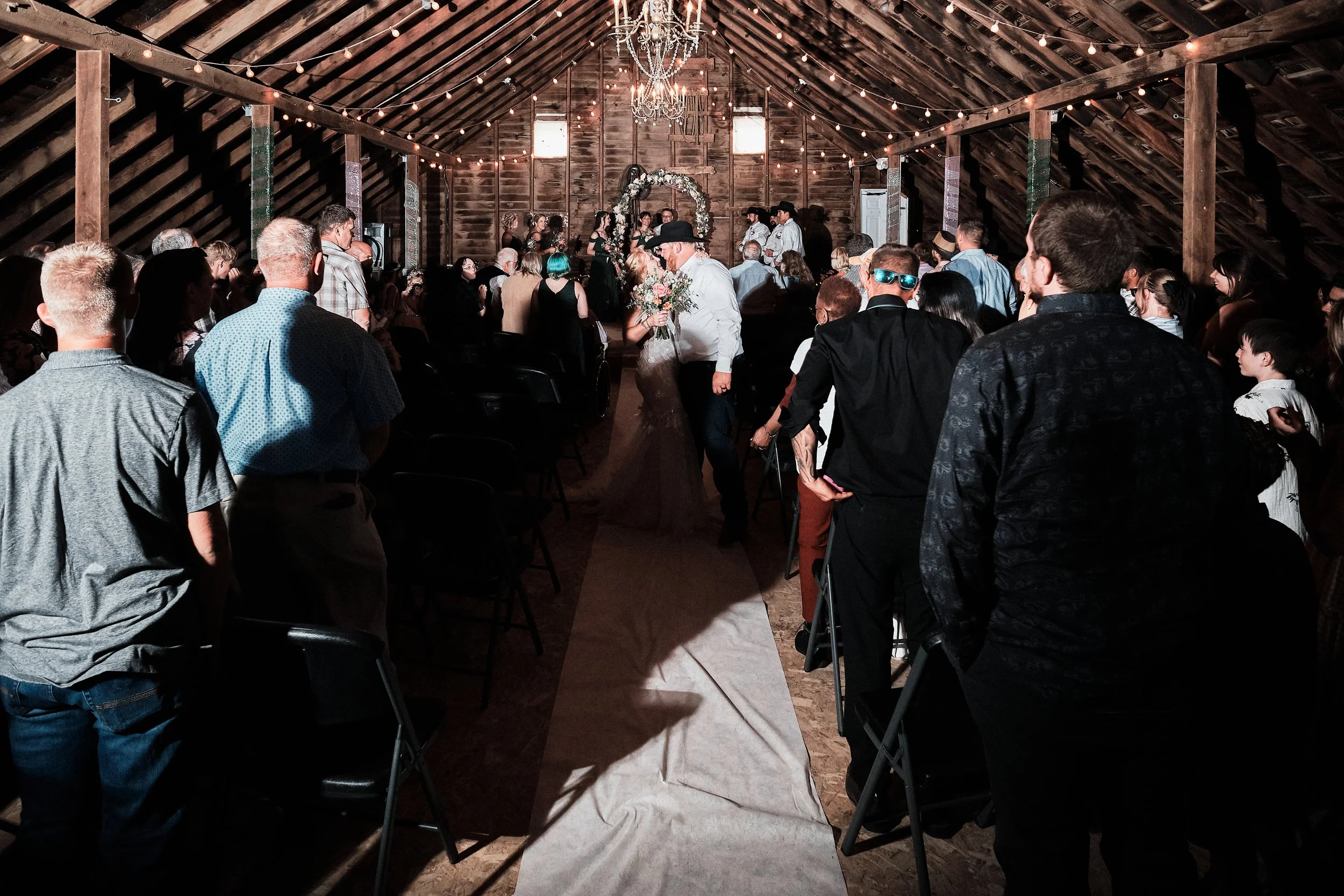 A wedding reception inside a rustic barn with string lights and a chandelier. People are gathered around a bride and groom walking down an aisle, with the bride holding a bouquet and the groom wearing a cowboy hat.
