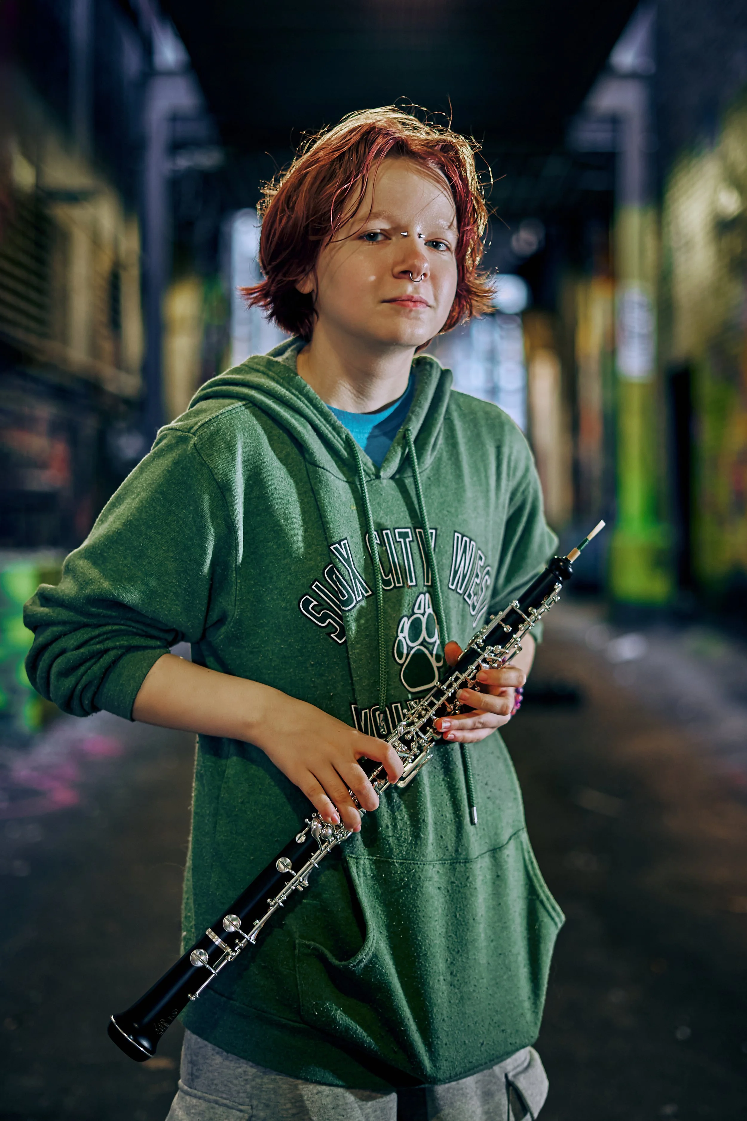 A young person with red hair holding a black clarinet in a dimly lit urban alleyway.