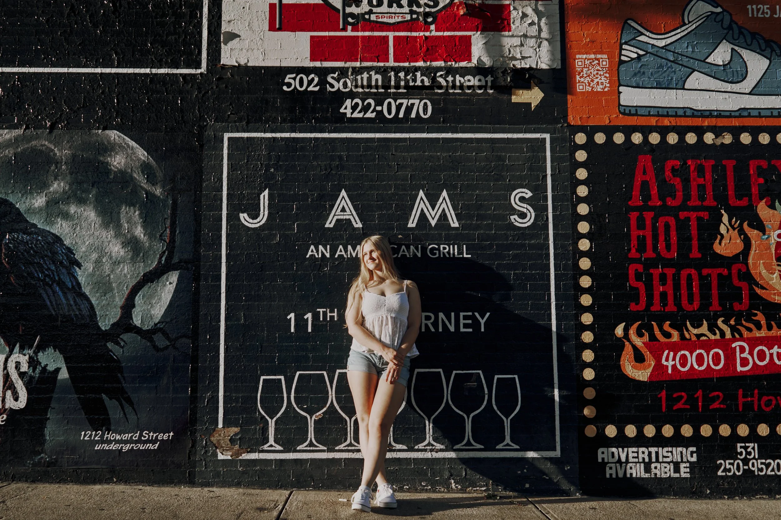 A young woman with long blonde hair in a white tank top and shorts stands in front of a large, colorful street mural on a brick wall, with her arms crossed and smiling.