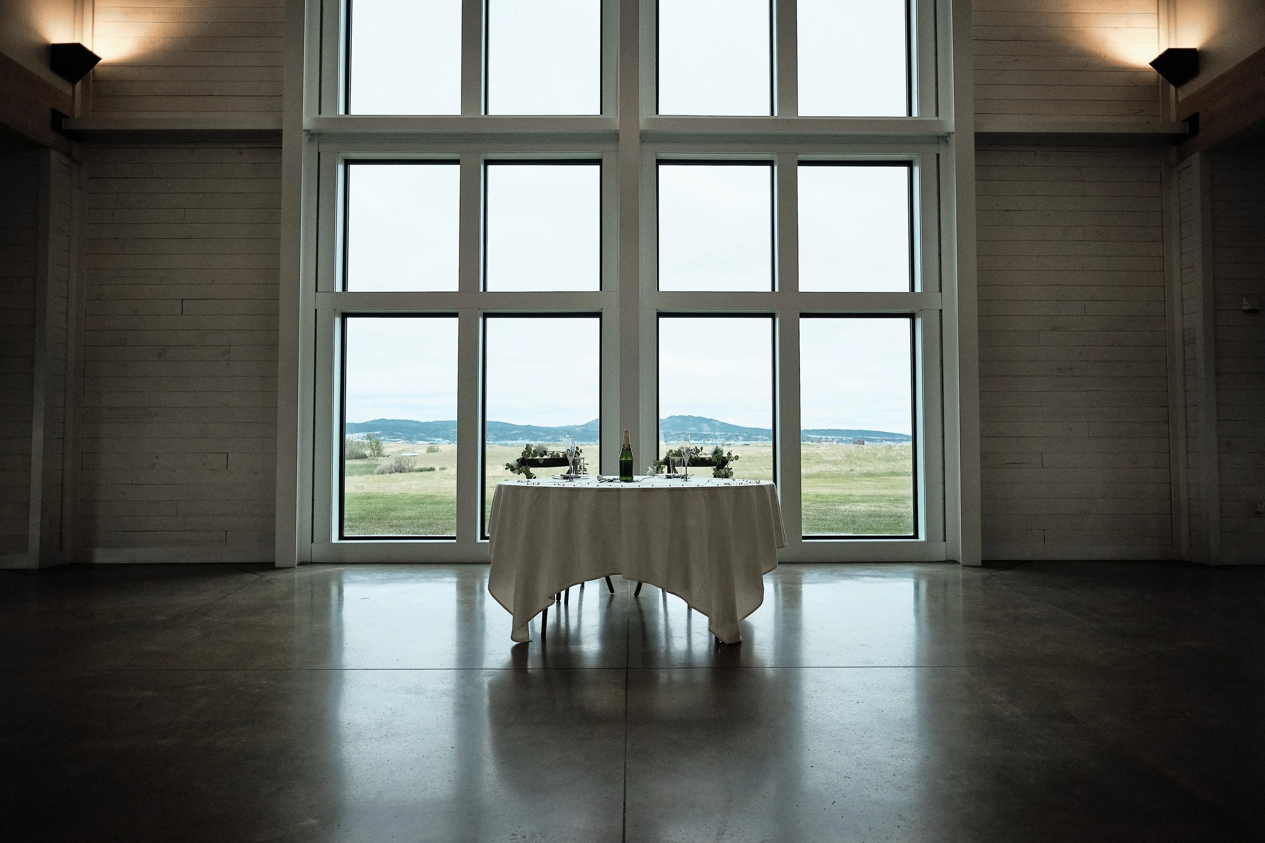 Interior of a modern room with large windows showing a scenic landscape. There is a round table with a white tablecloth and a bottle of champagne, set for a meal.