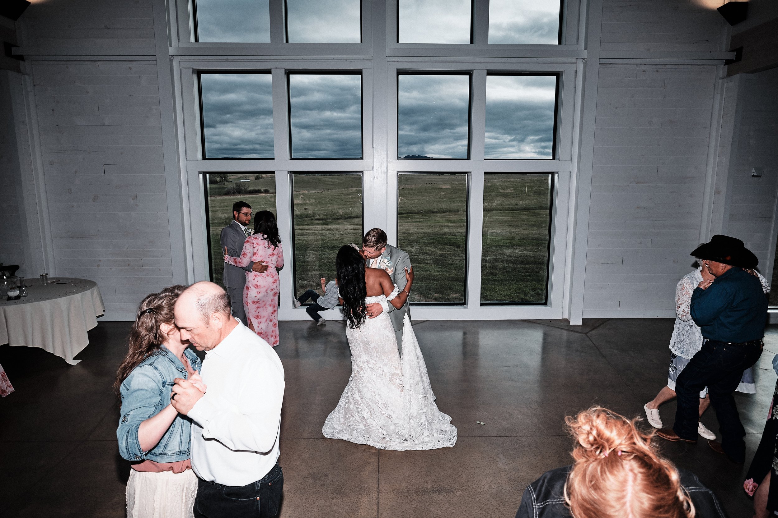A bride and groom dancing at their wedding reception in a large room with a tall window showing a cloudy outdoor sky. Guests are gathered around, some dancing, some seated.