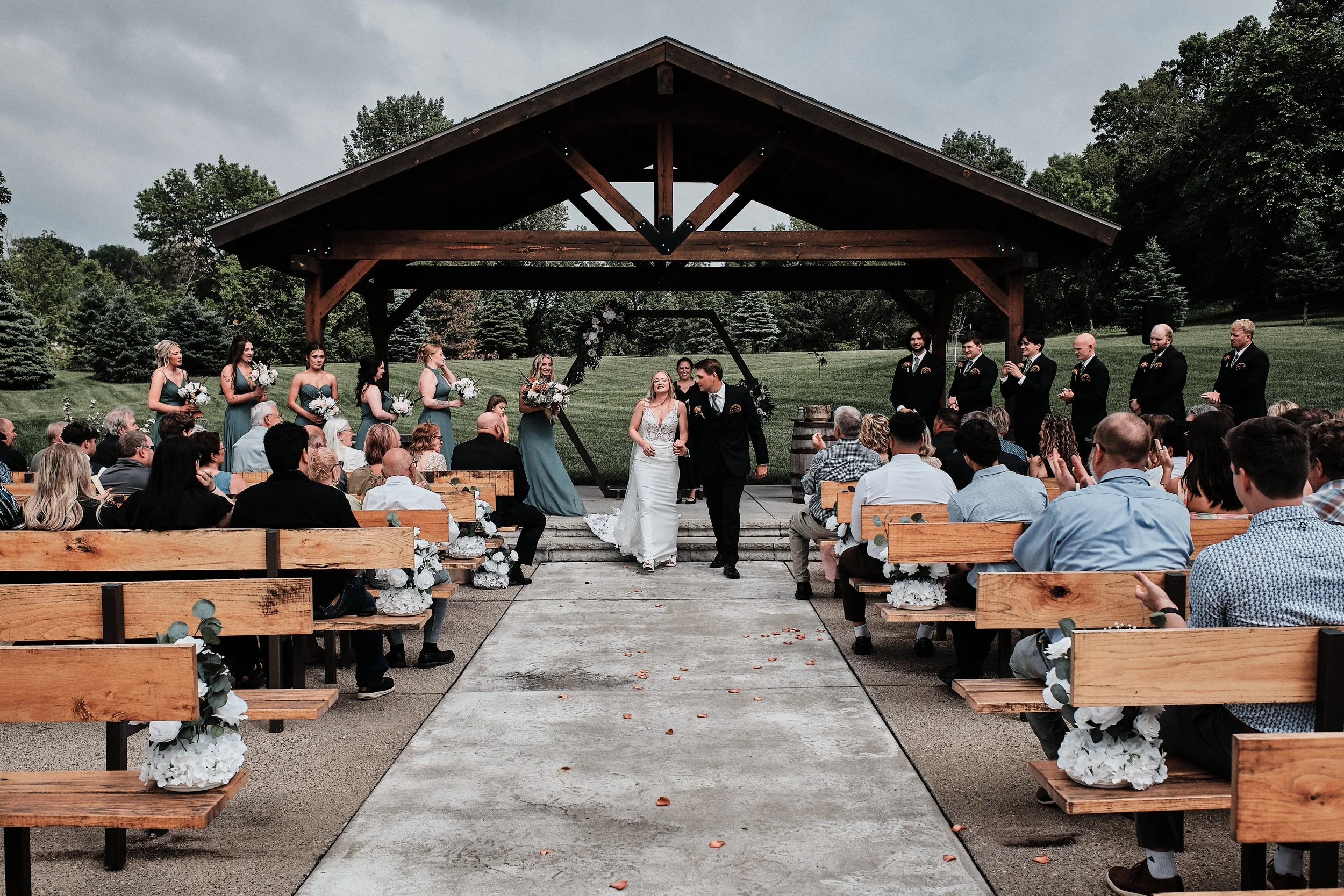 Outdoor wedding ceremony with a bride and groom standing under a wooden arch, surrounded by bridesmaids and groomsmen, with seated guests and a backdrop of trees.